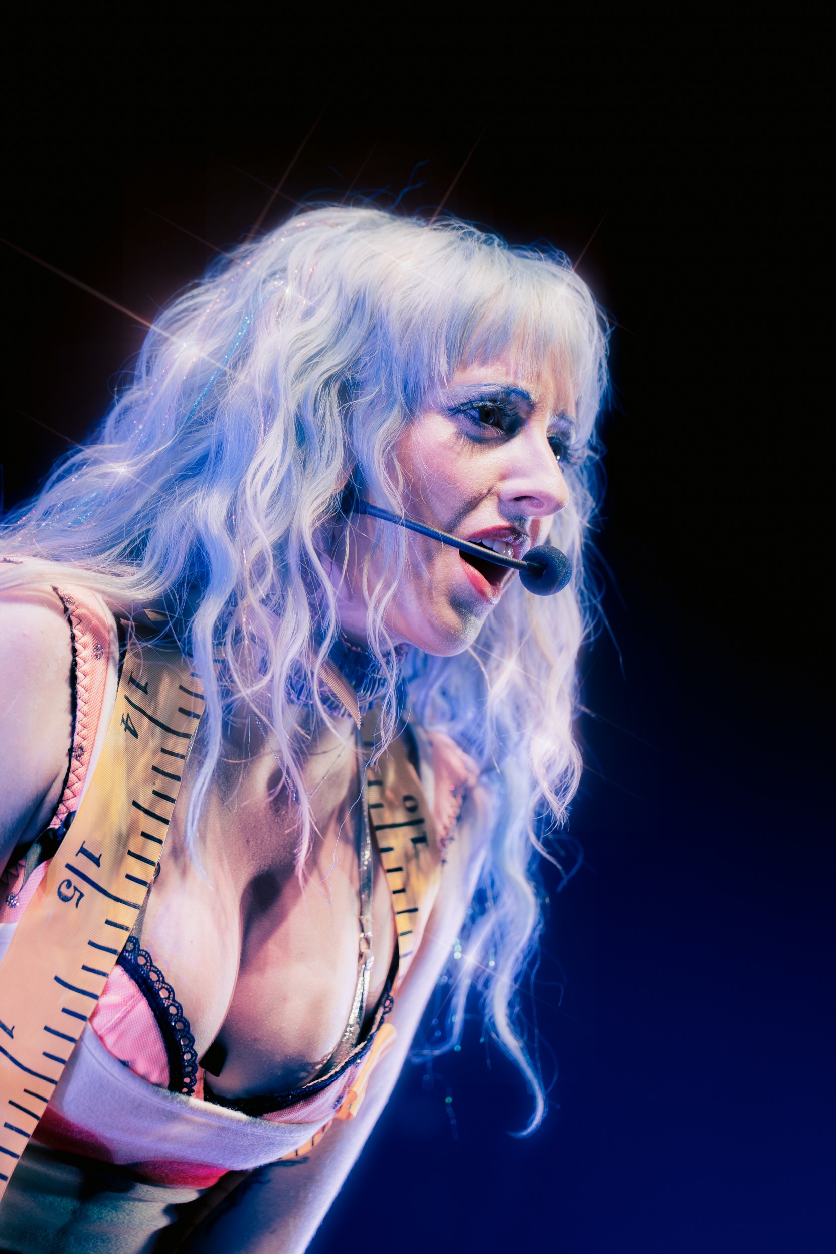 Performer with long hair and mic on stage, wearing creative clothing.