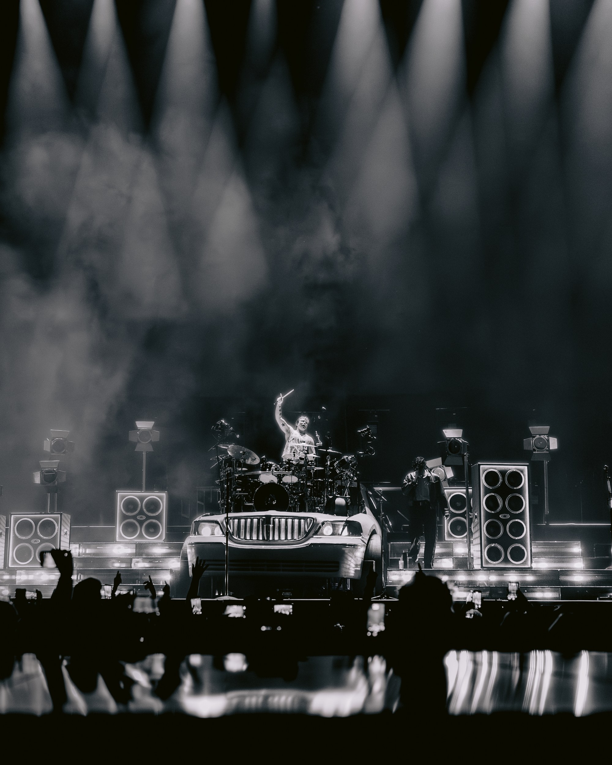 Dramatic live concert scene with drummer on elevated car stage, intense lighting effects.