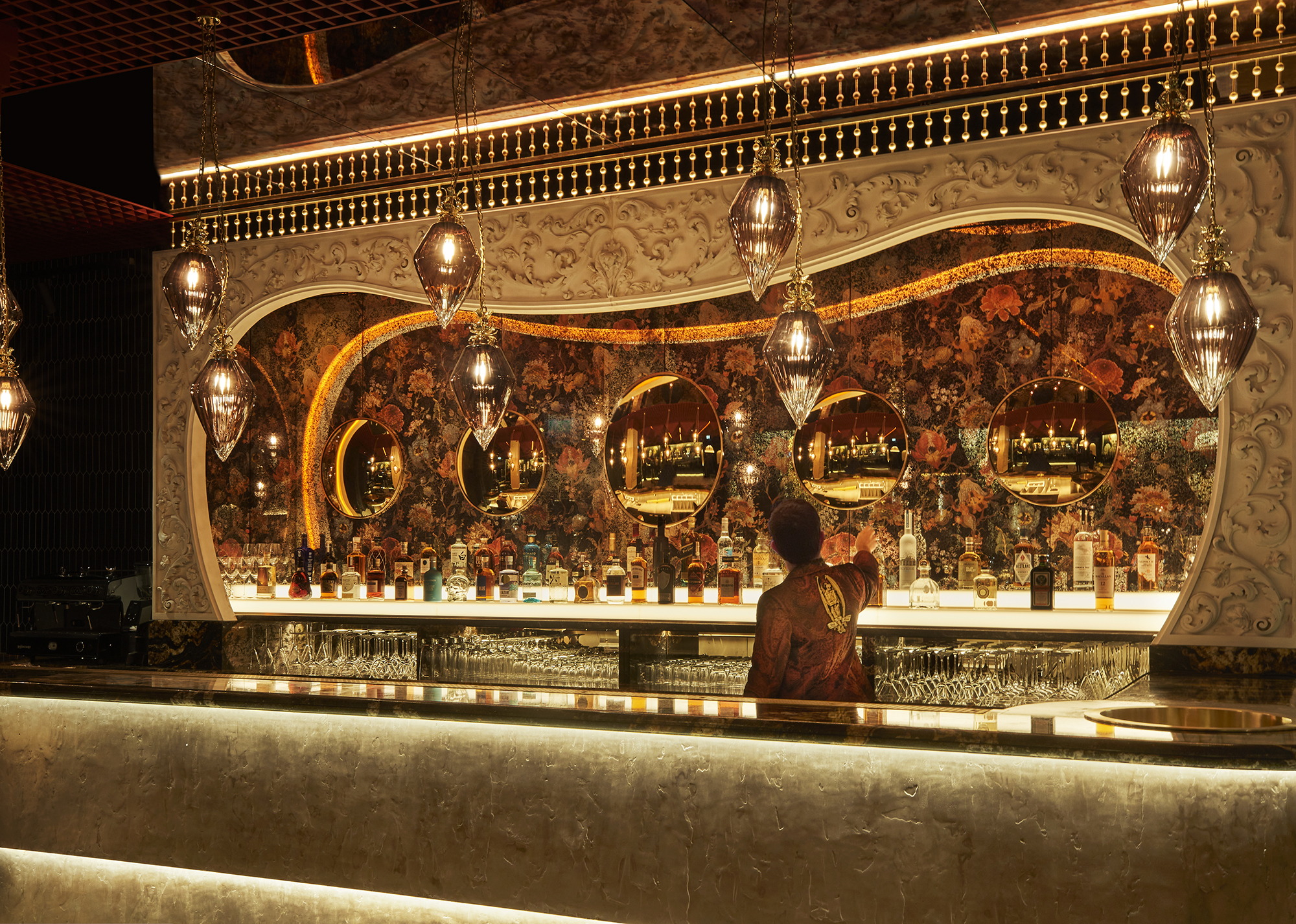 Elegant bar with floral wallpaper, hanging lights, and a bartender selecting drinks.