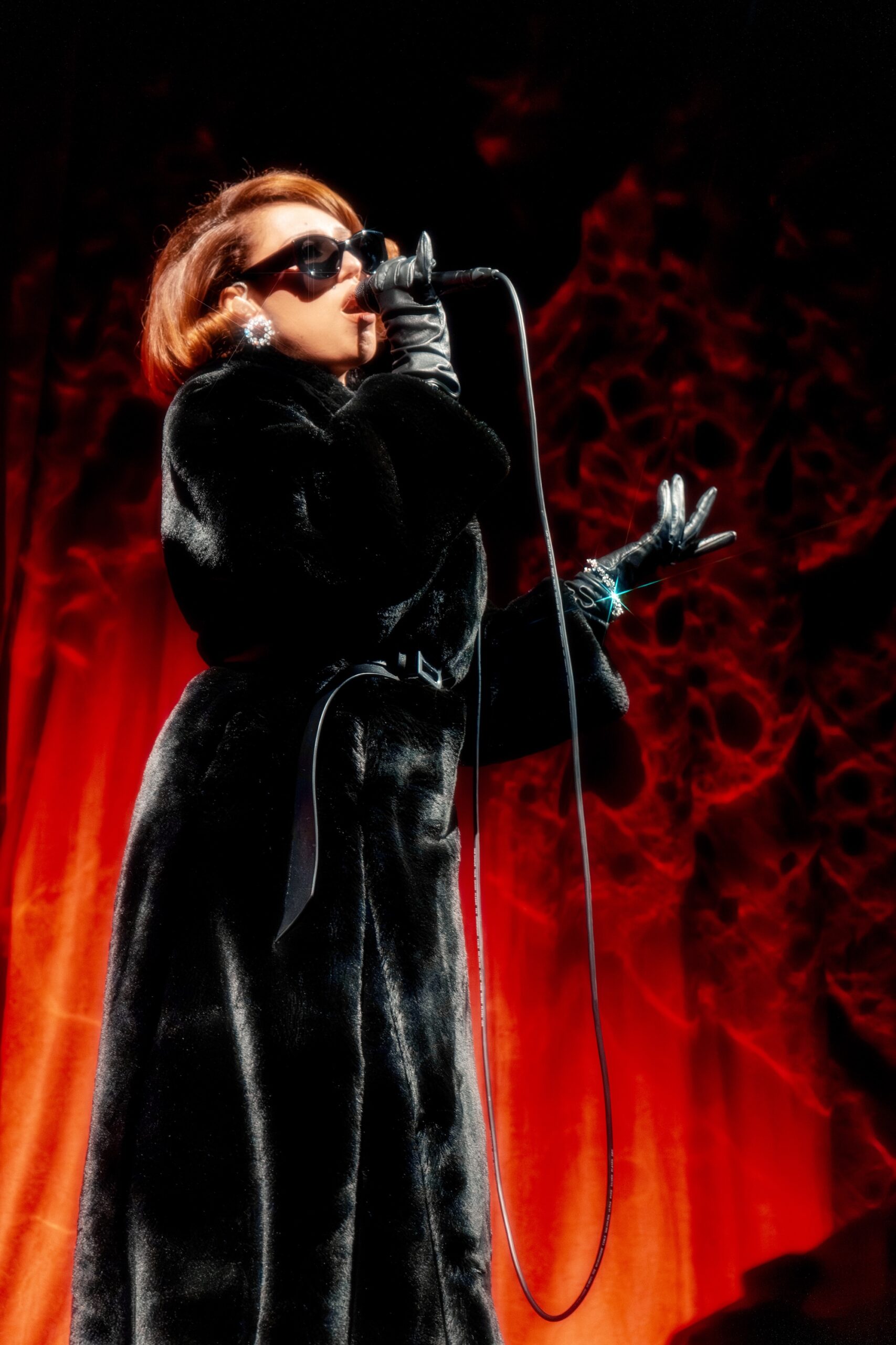 Singer in black coat and gloves performs passionately on stage with red backdrop.
