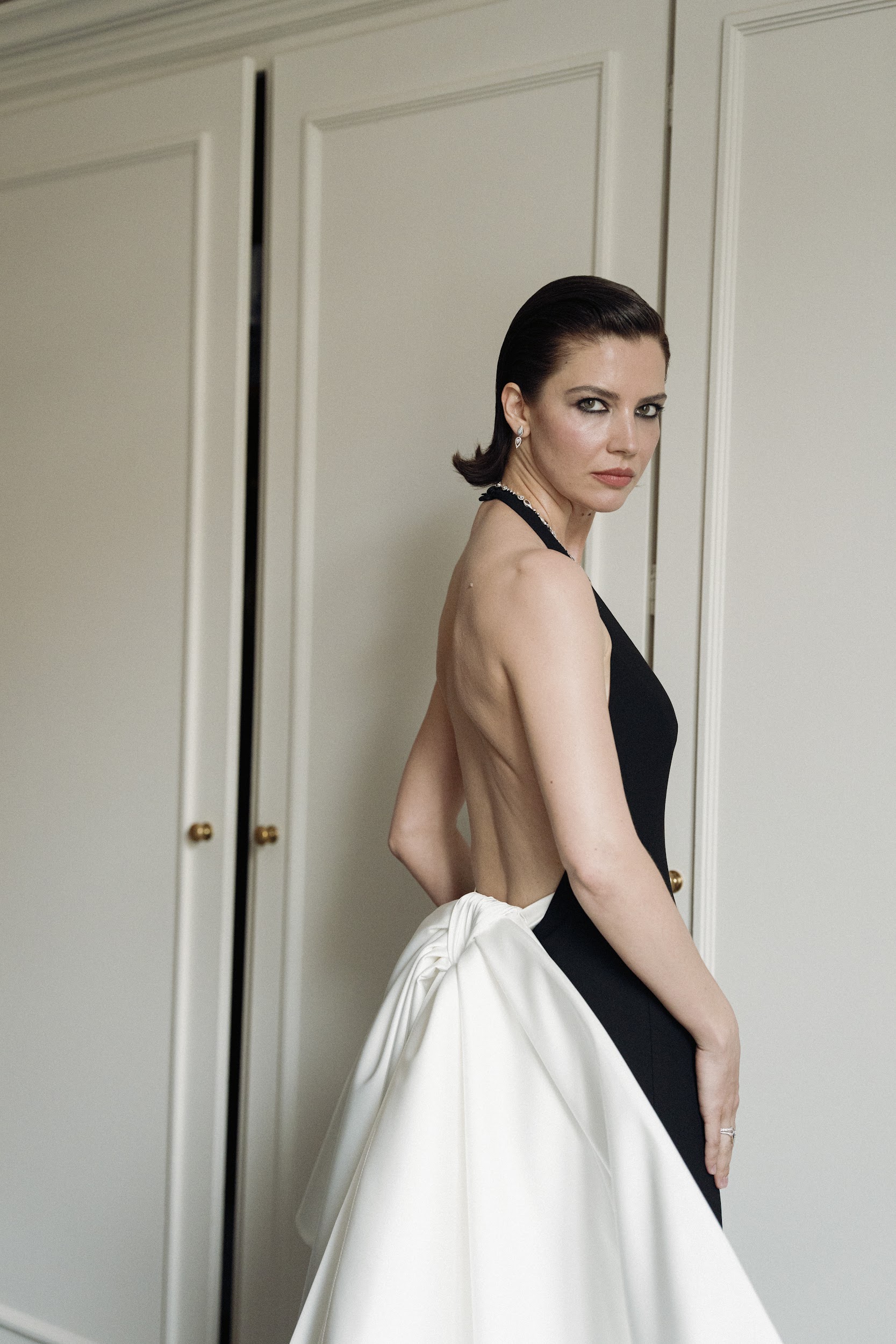 Elegant woman in a backless black gown with white bow detail, standing by a door.