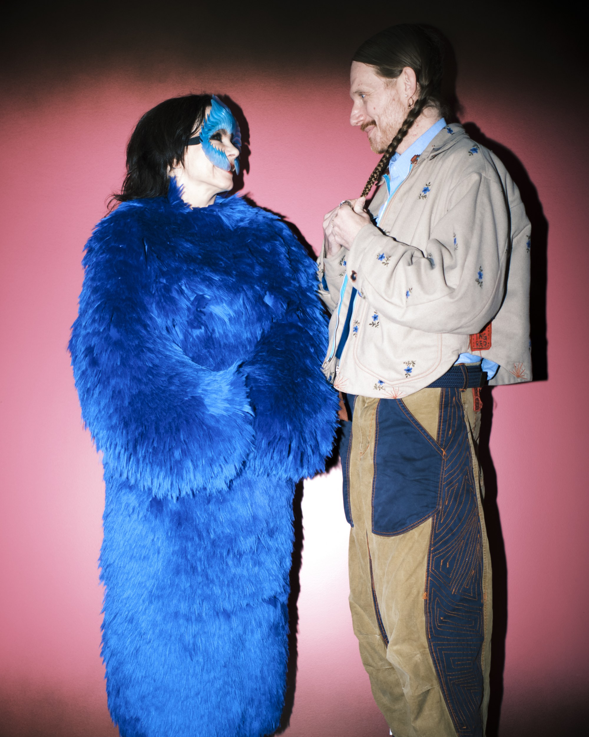 Person in blue feather costume faces another person against a pink background.