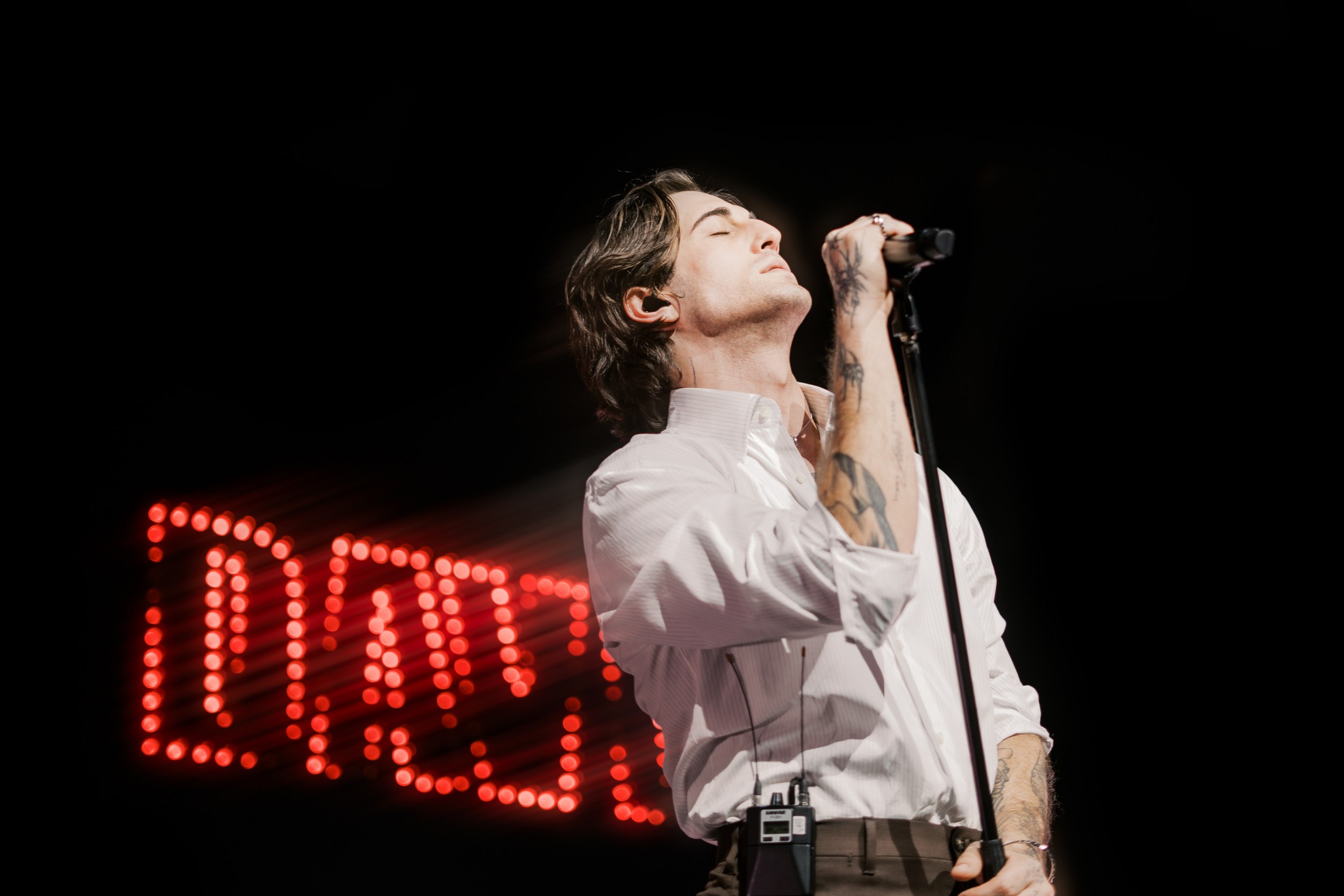 Singer passionately performing on stage with red neon lights in the background.