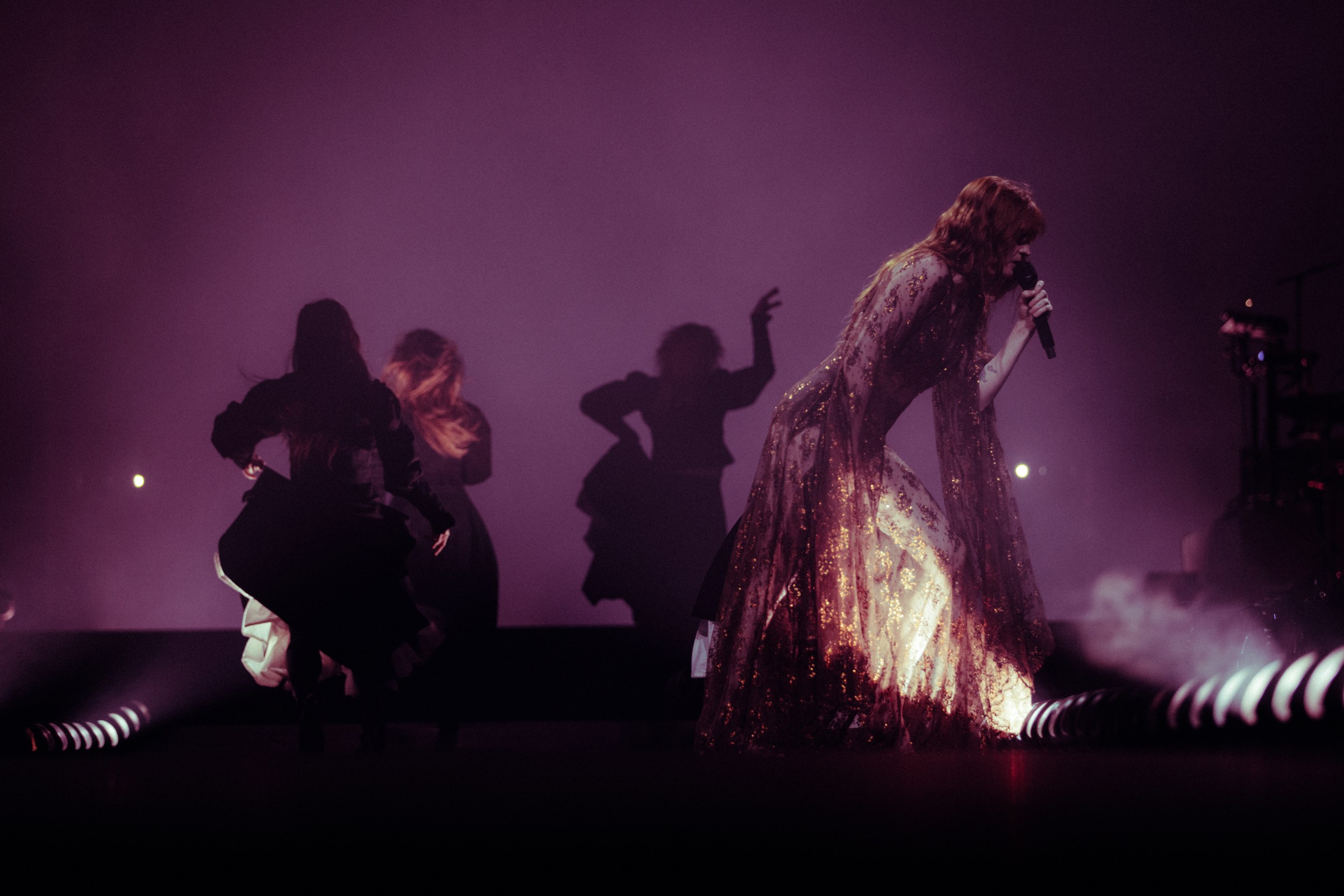 Singer performing dramatically onstage with dancers silhouetted in purple lighting.