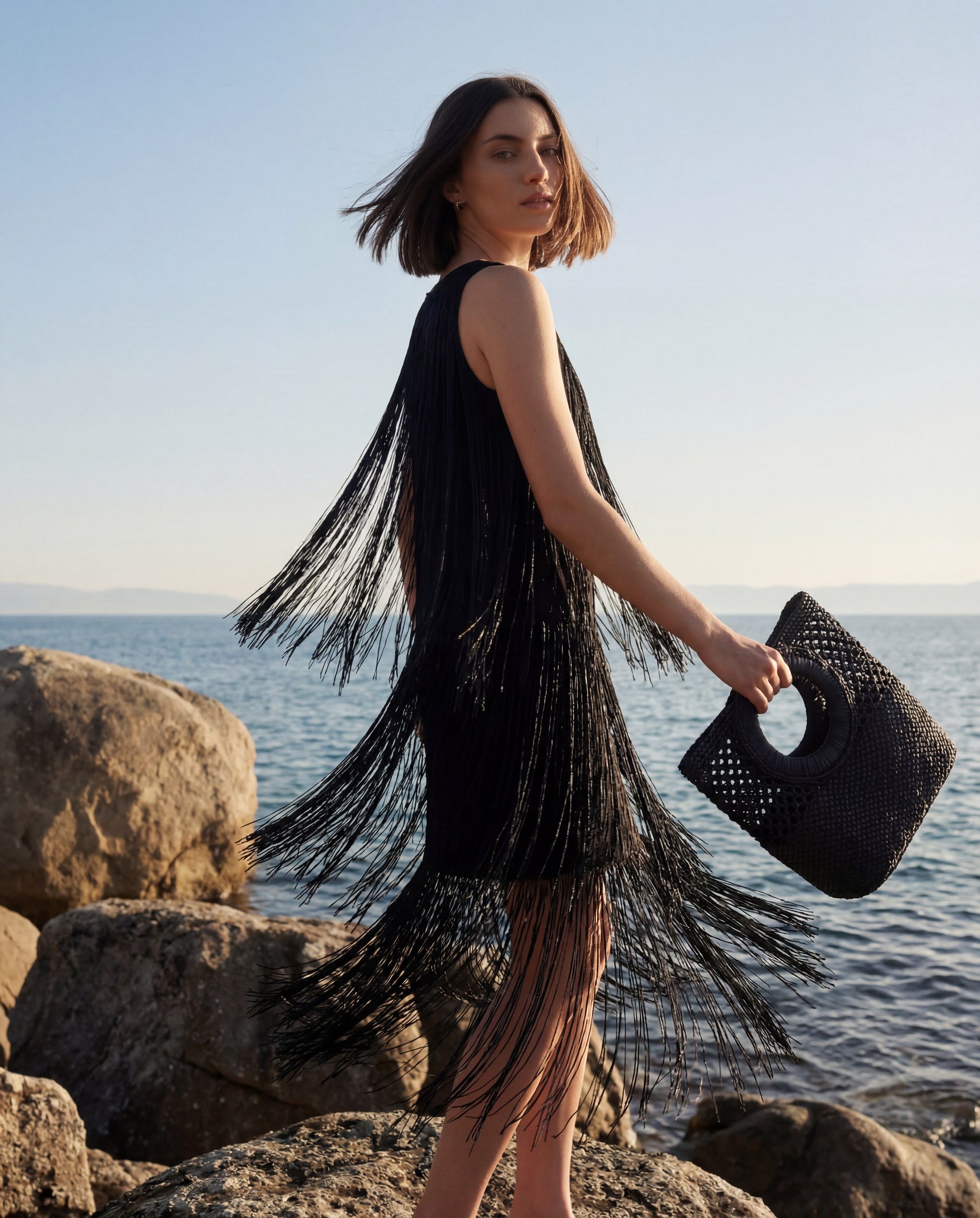 Woman in black fringe dress by ocean with woven handbag.