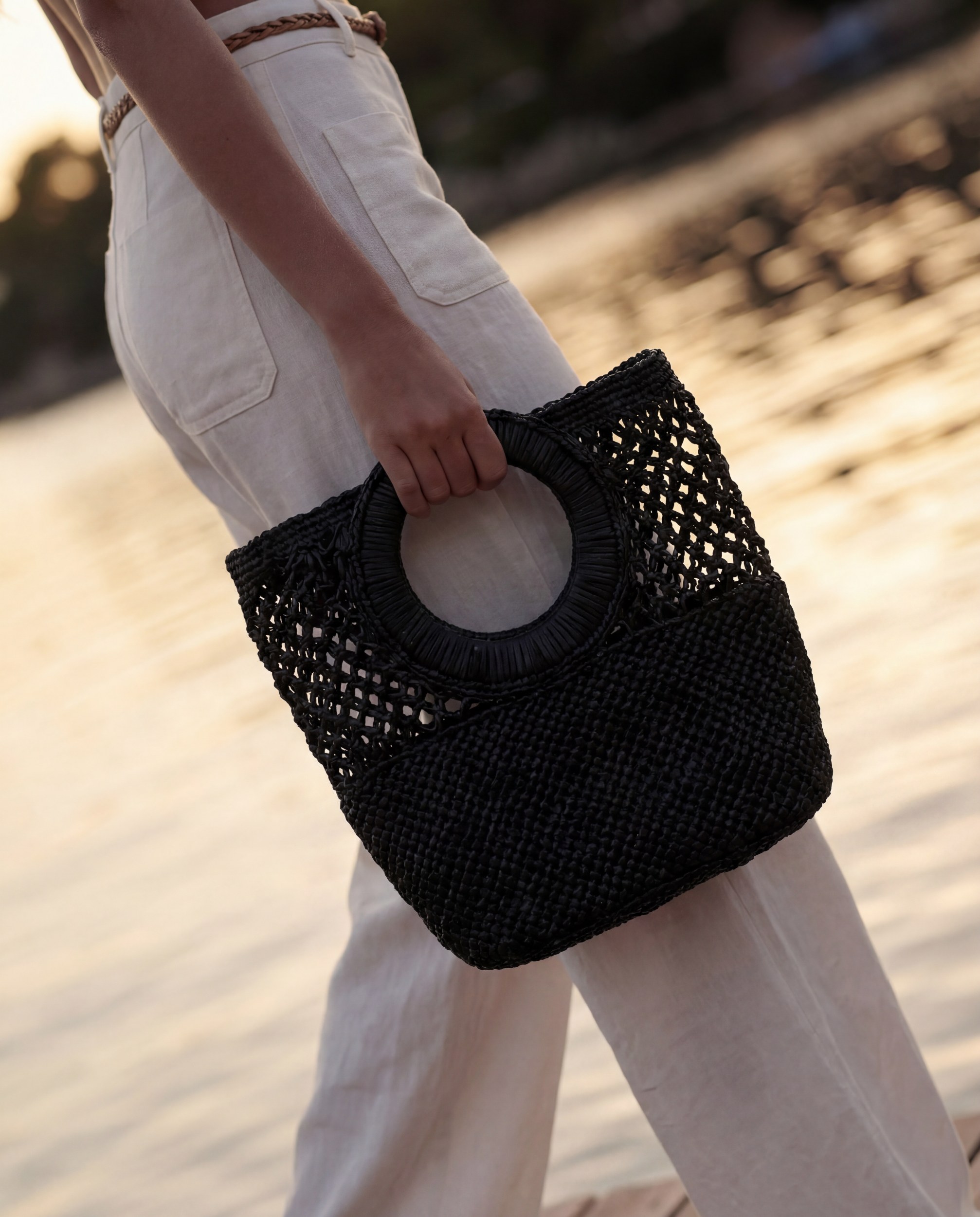 Stylish woven black handbag paired with light trousers by the water at sunset.