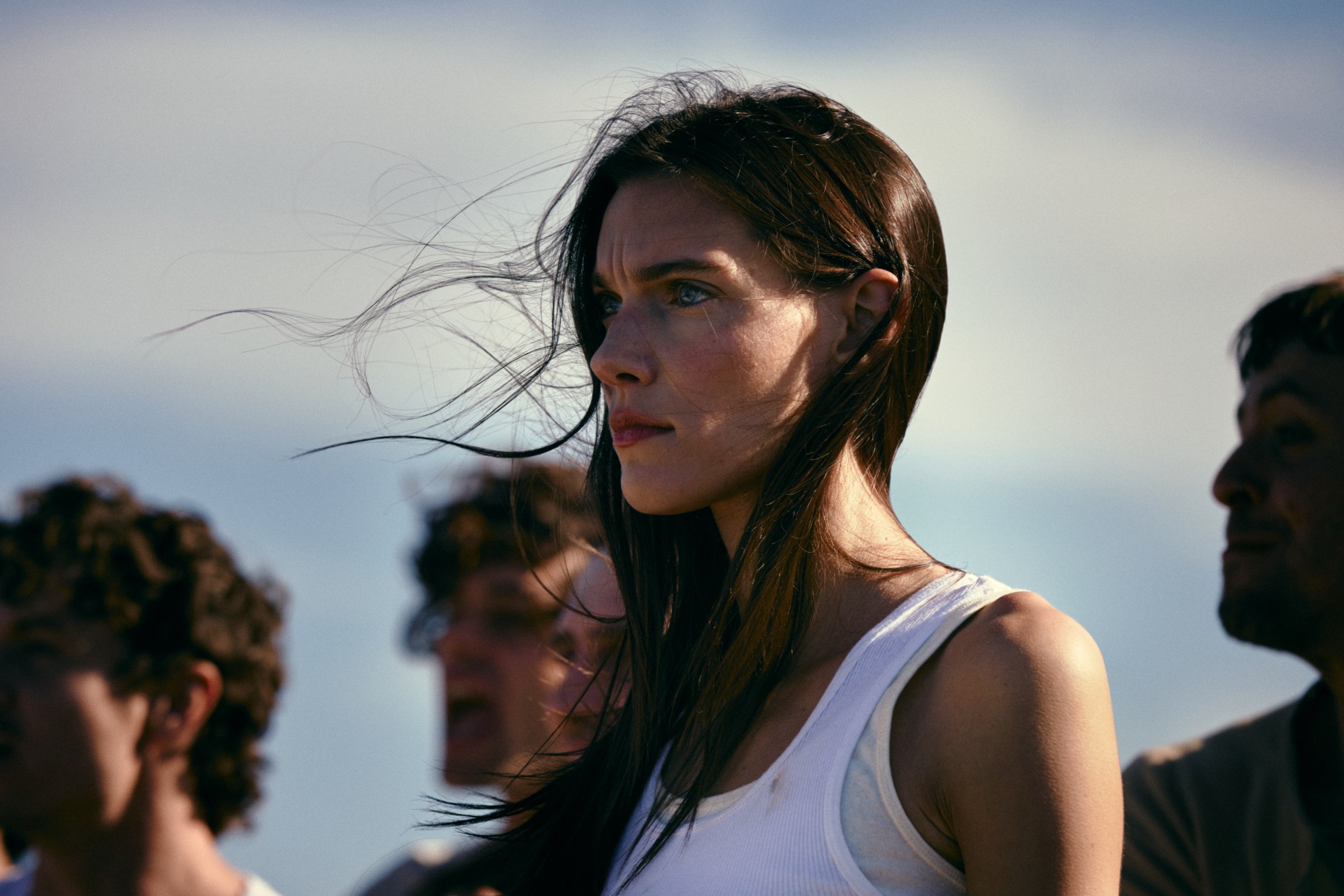 Woman gazing into distance, wind in hair, wearing white top, against sky background.