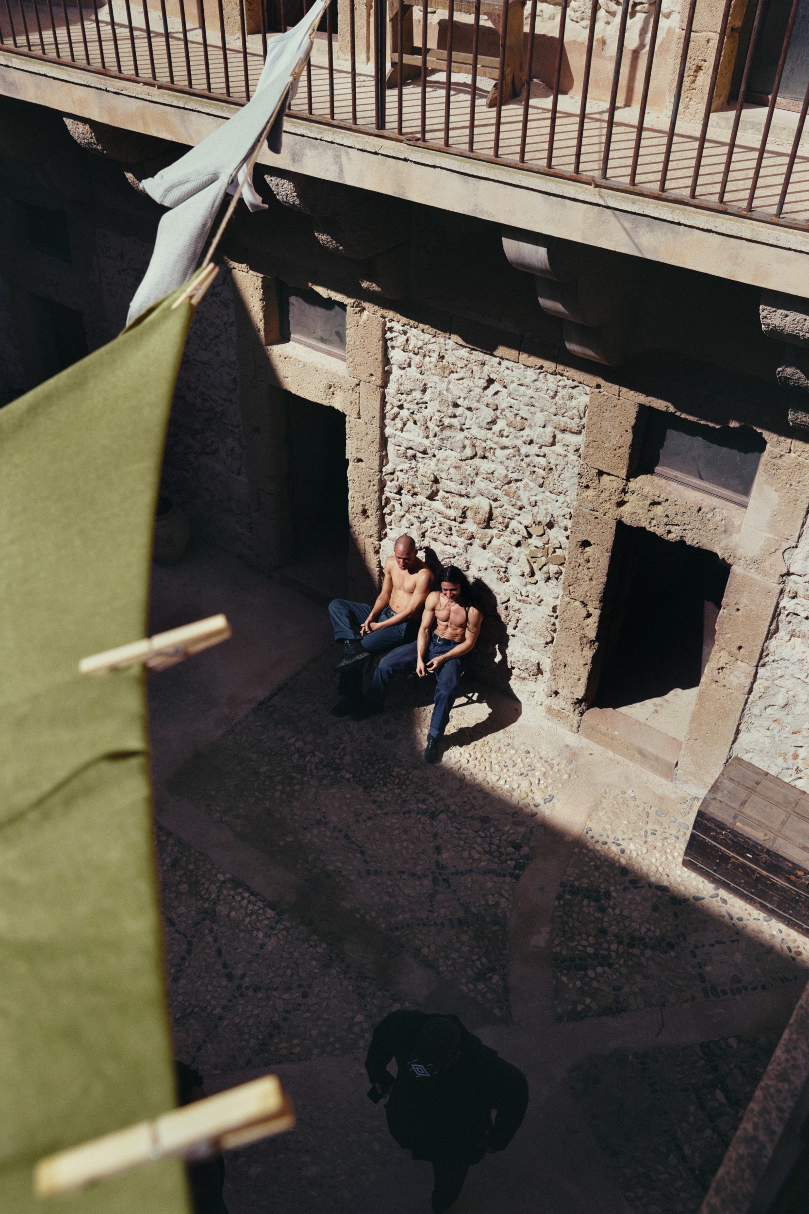 Two men sitting shirtless in a sunlit courtyard with hanging laundry.