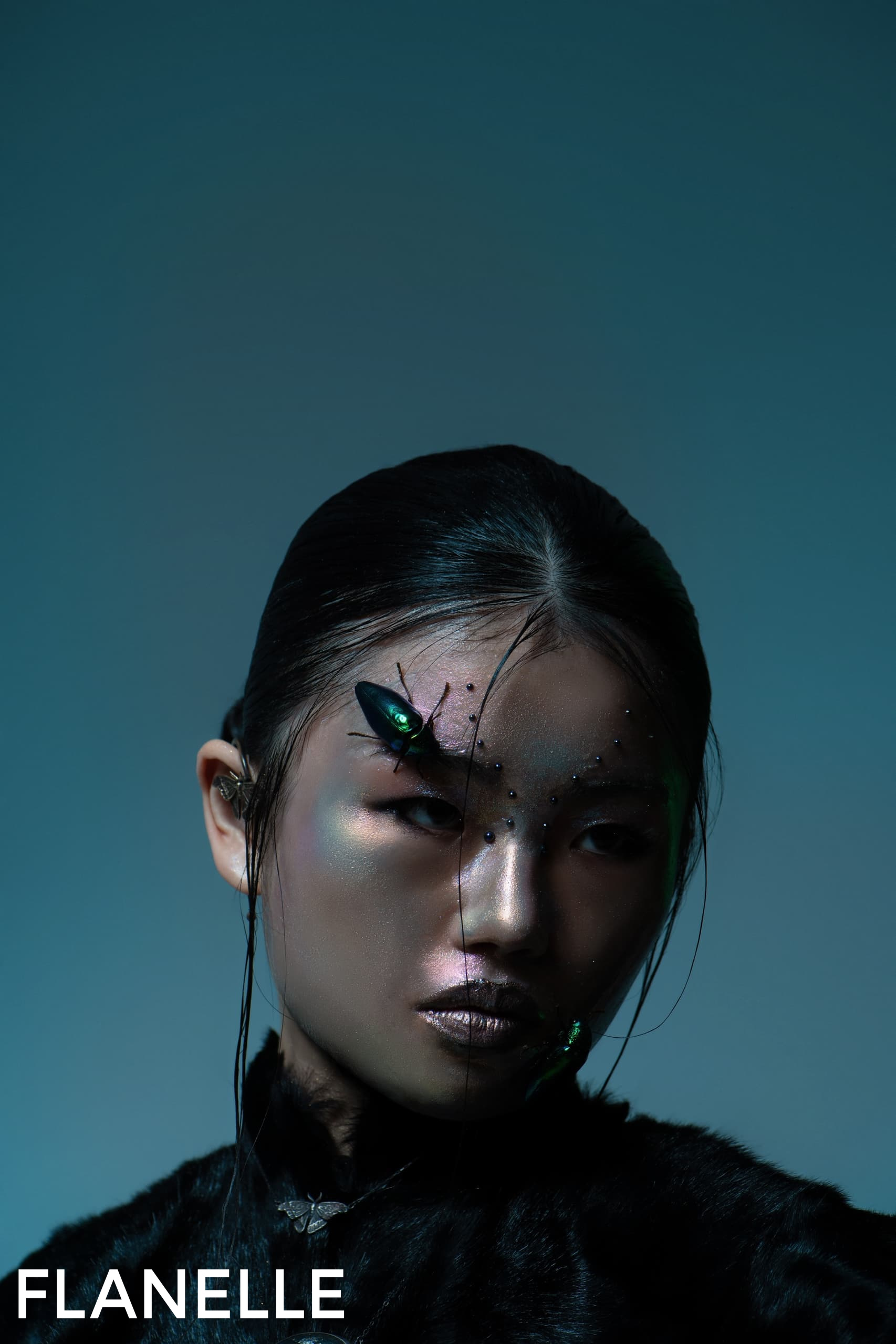 Model with futuristic makeup and insect accessory, dark background.