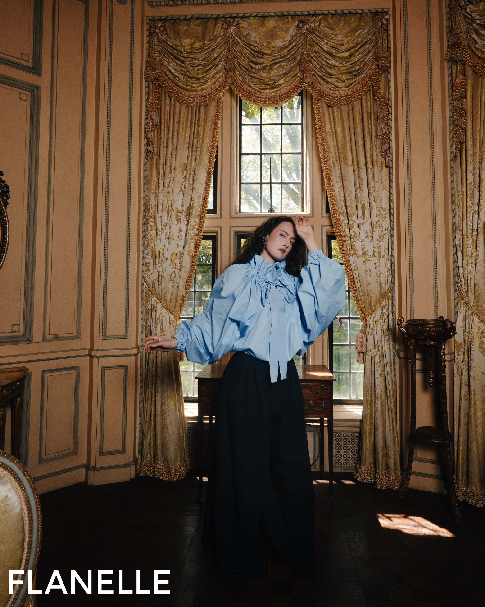 Fashion model poses elegantly in luxurious room with draped curtains.