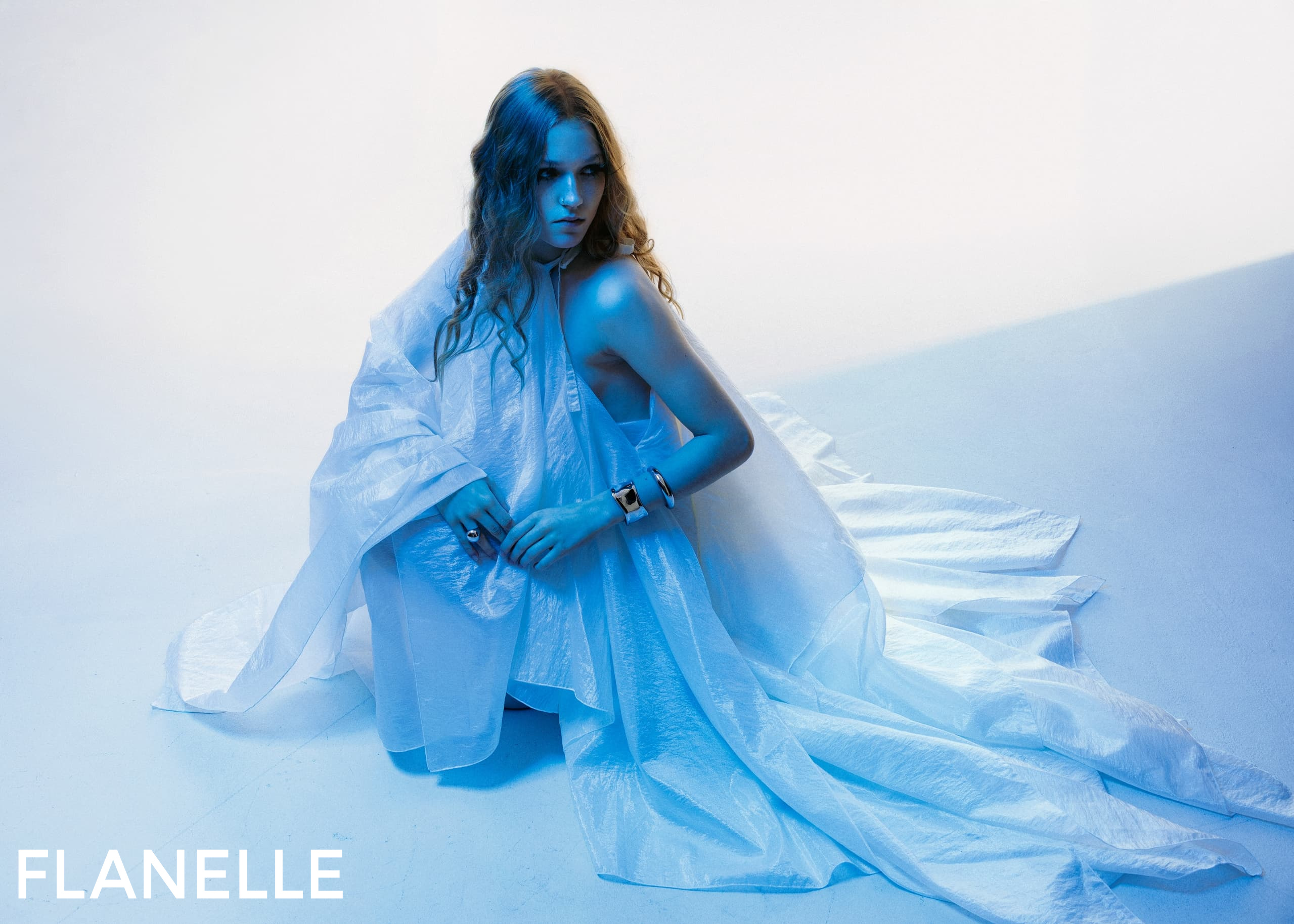 Model in flowing blue dress, seated in dramatic lighting, Flanelle magazine shoot.