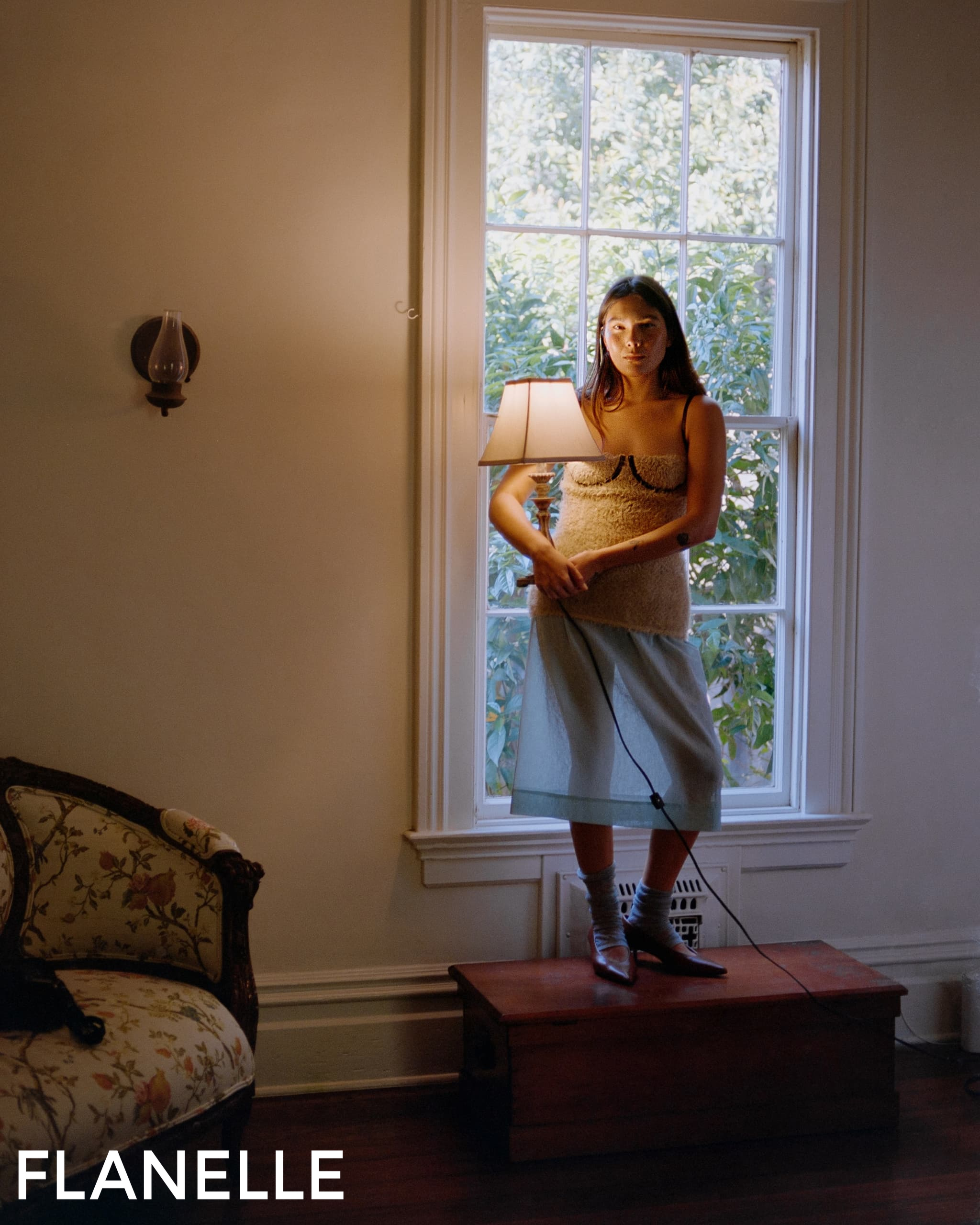 Woman standing by window in vintage room, wearing stylish sweater and skirt.