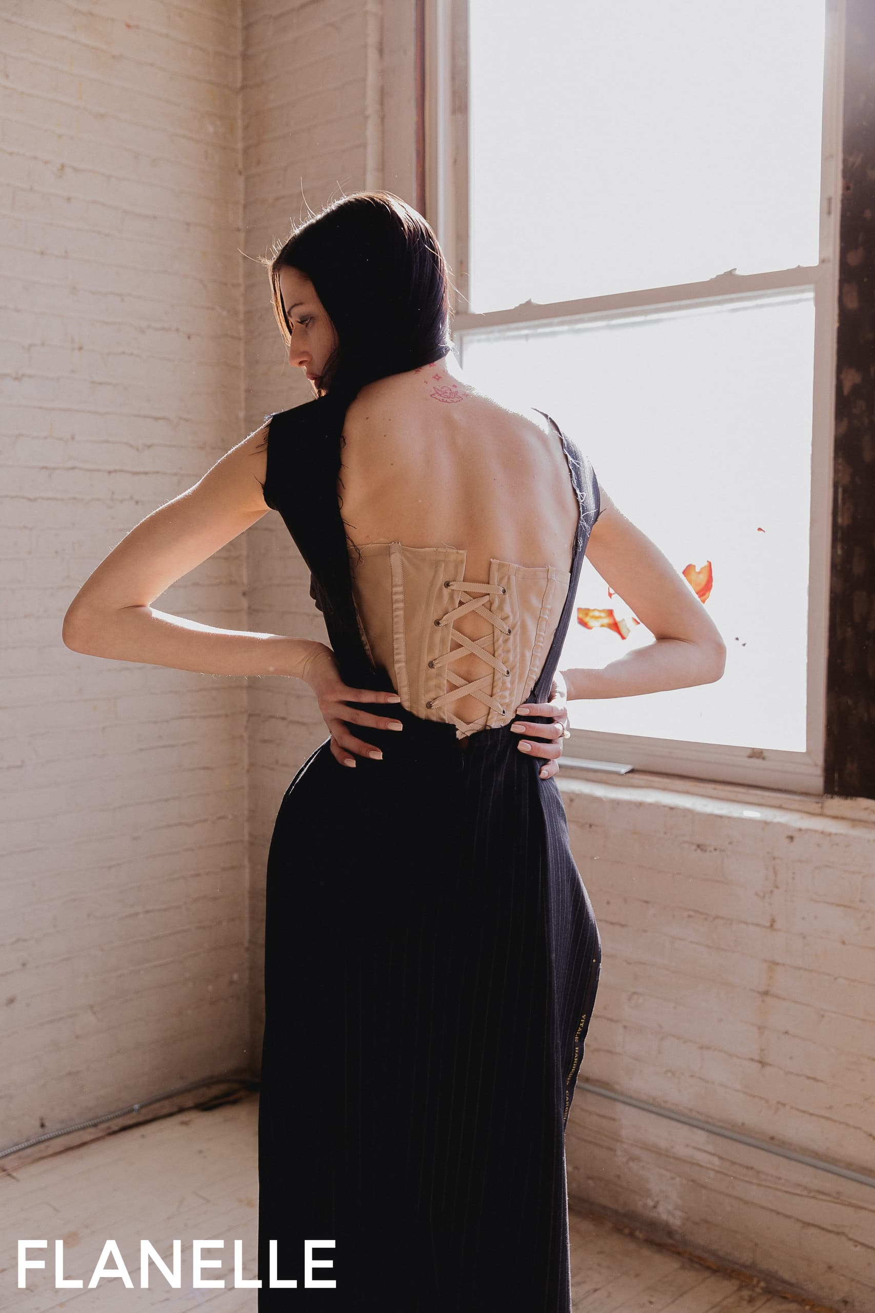 Fashion model in black gown with lace-up back detail by window.