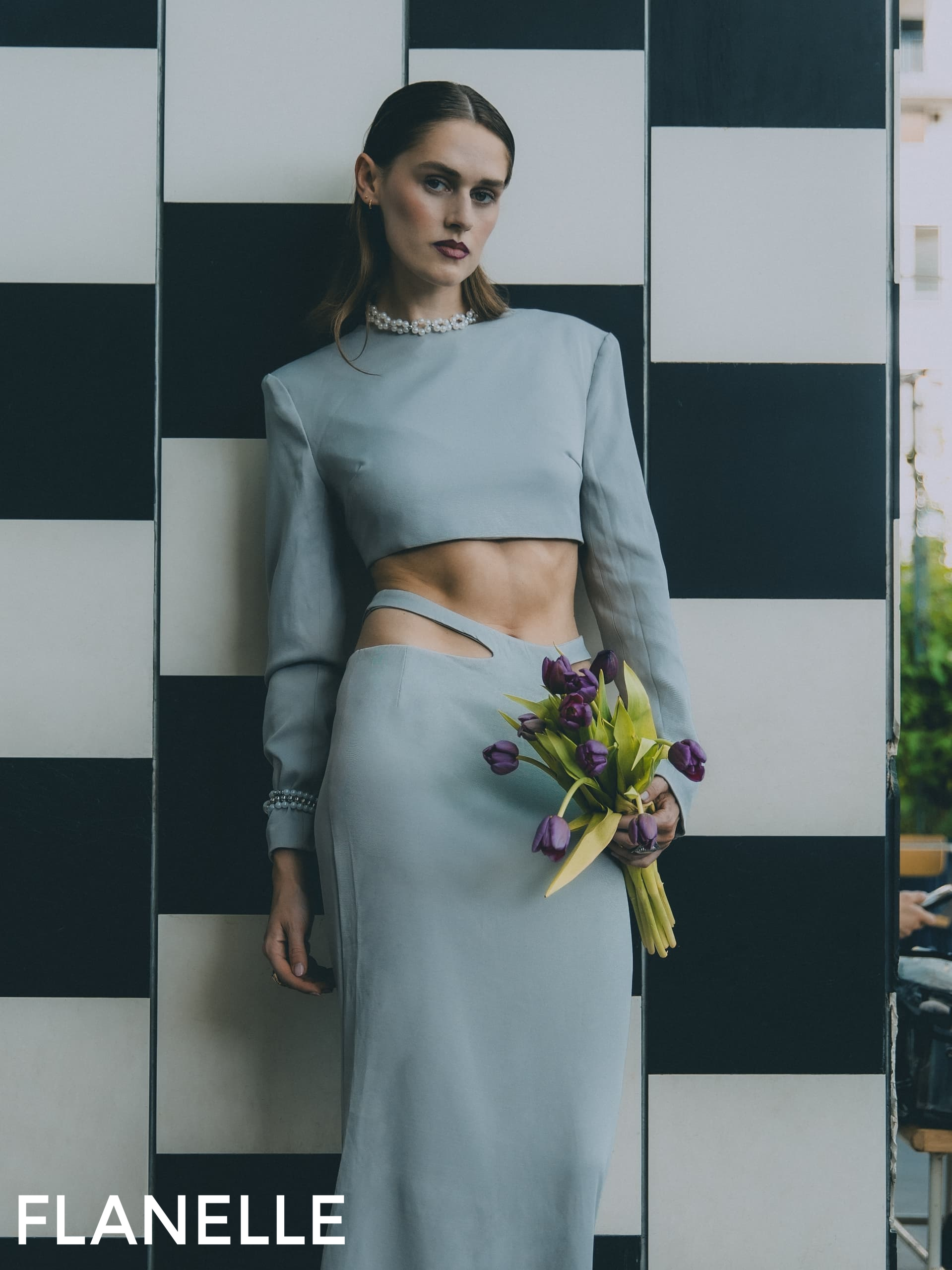 Elegant woman in chic attire holding purple flowers against a checkered backdrop.