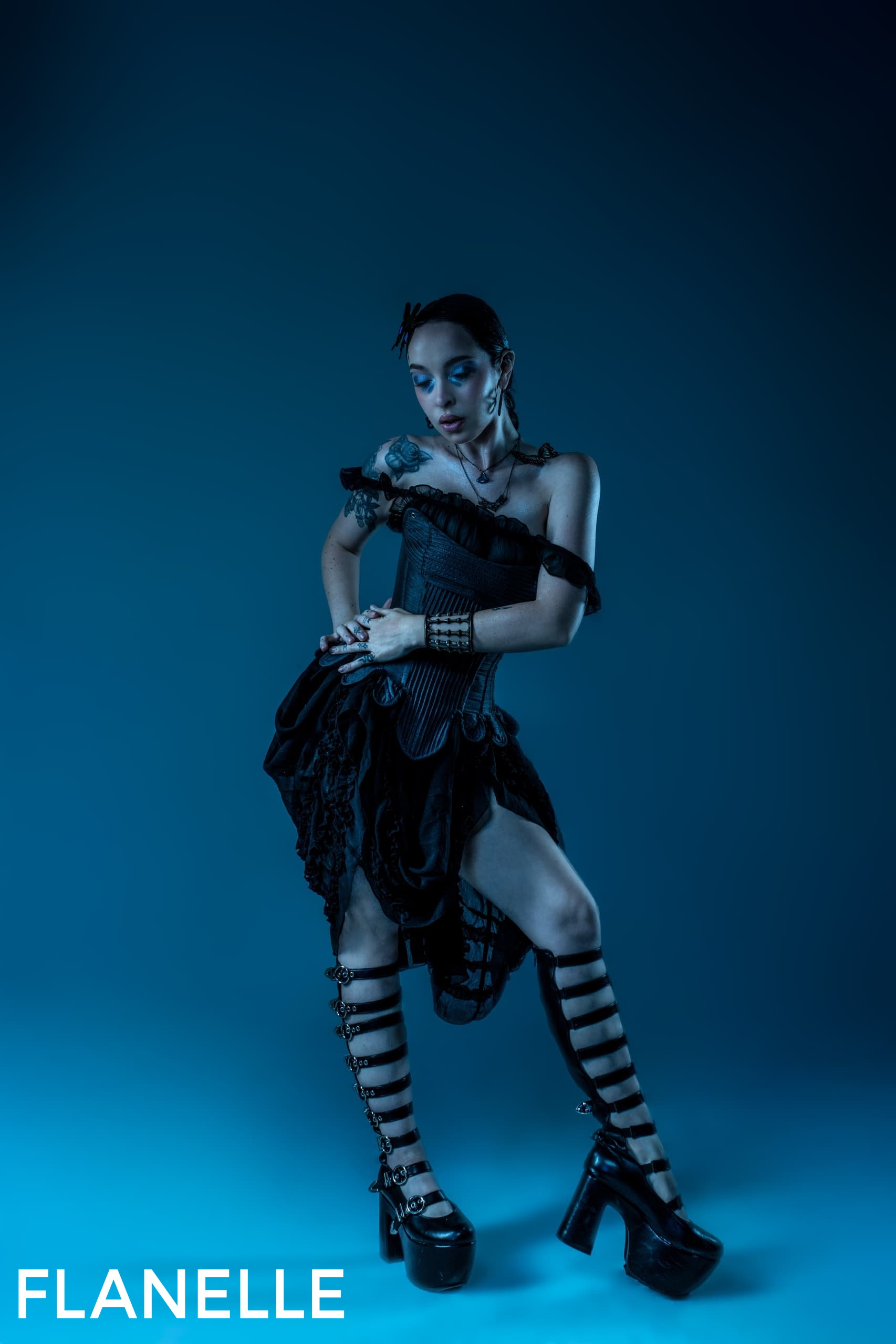 Gothic fashion model in corset dress and platform heels against dark blue background.