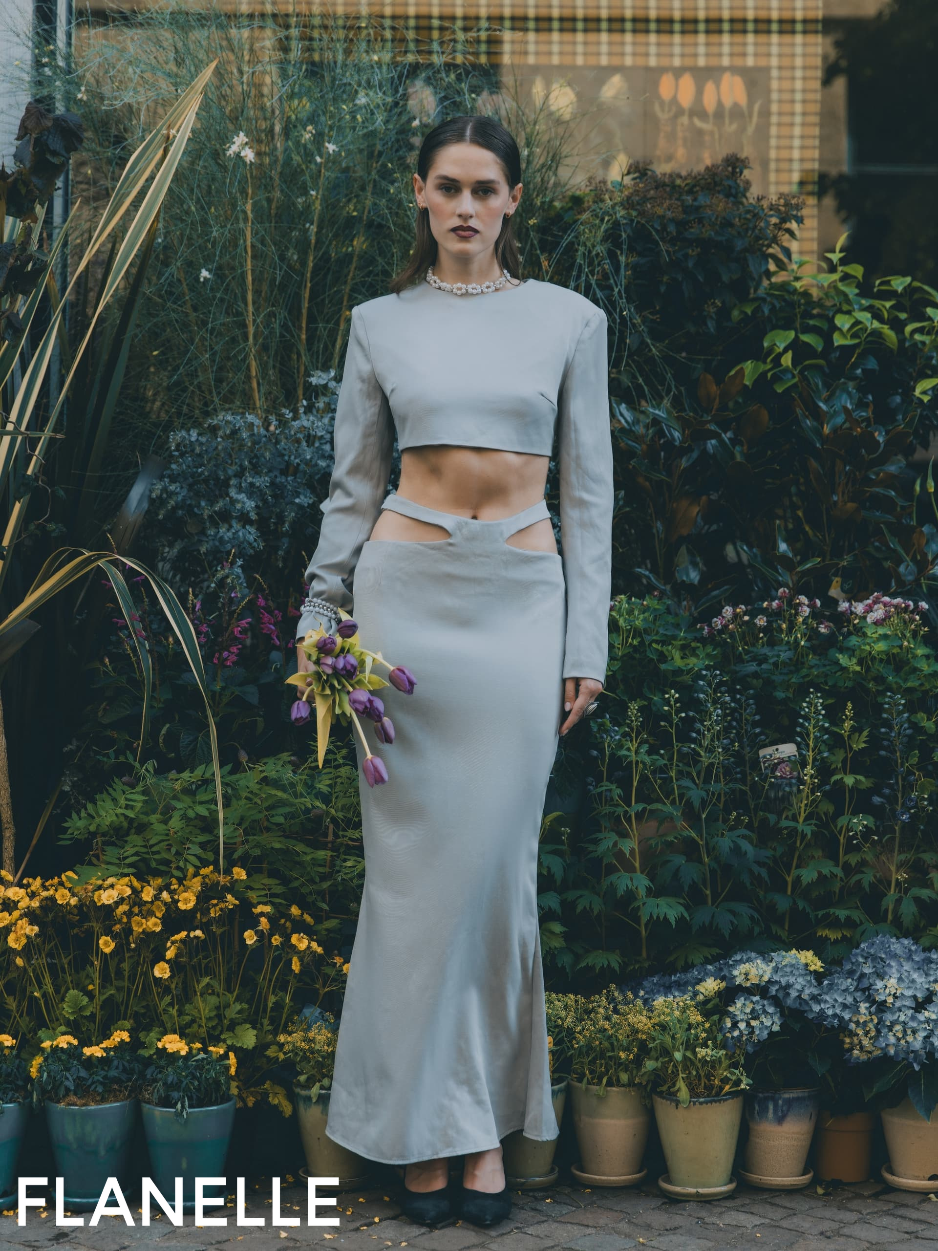 Fashion model in a chic gown holding purple tulips in a lush garden setting.
