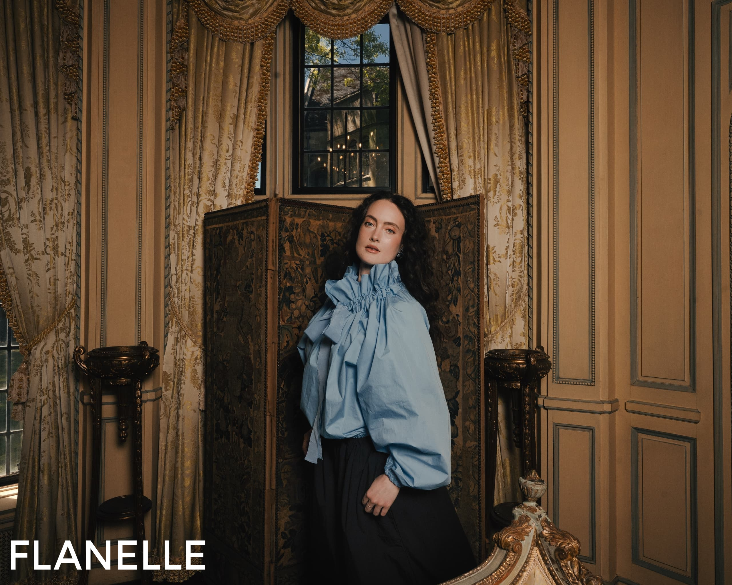 Elegant woman in blue blouse poses in luxurious, vintage-inspired room.