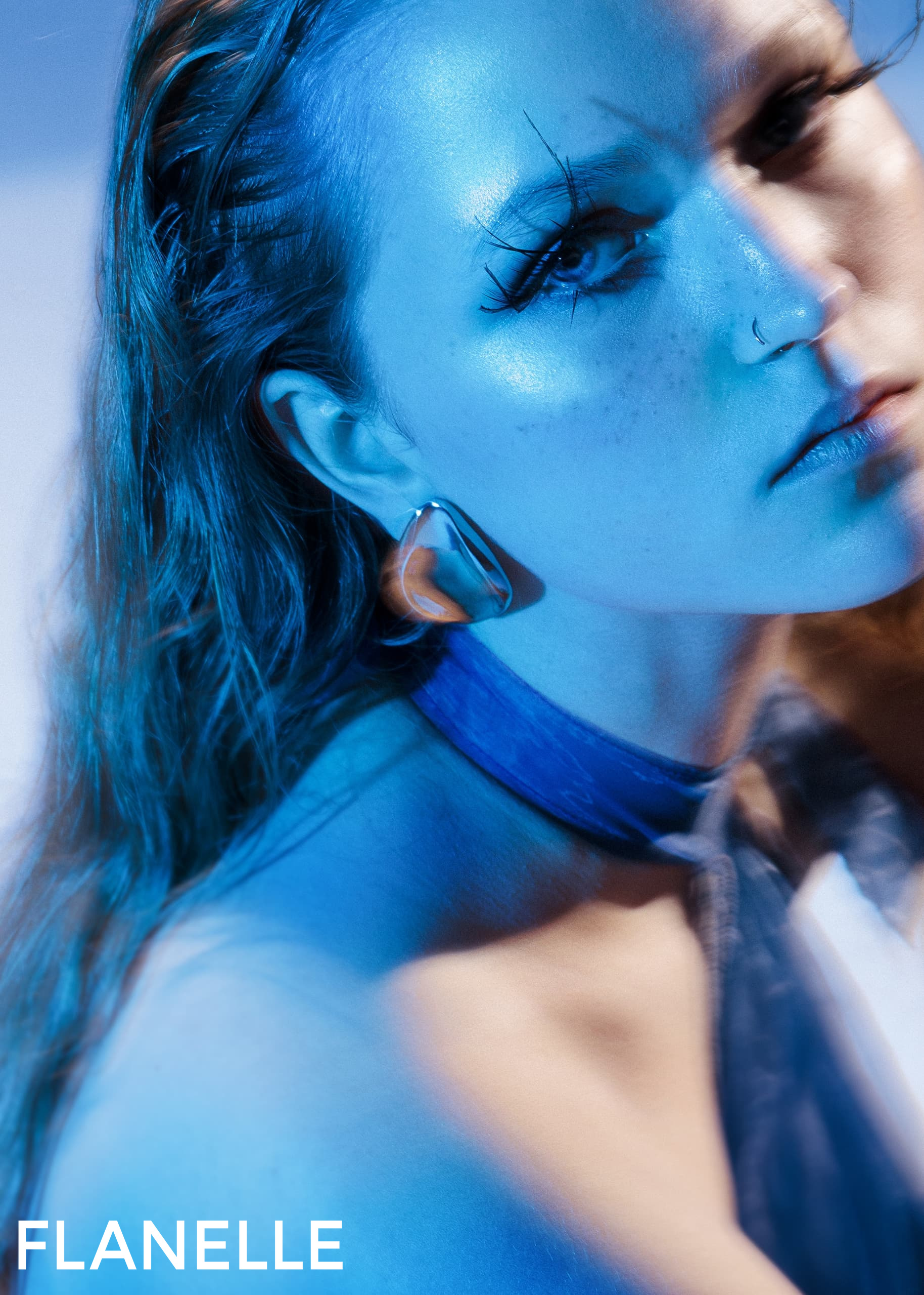 Fashion model in blue light with striking makeup and bold earring, Flanelle magazine.