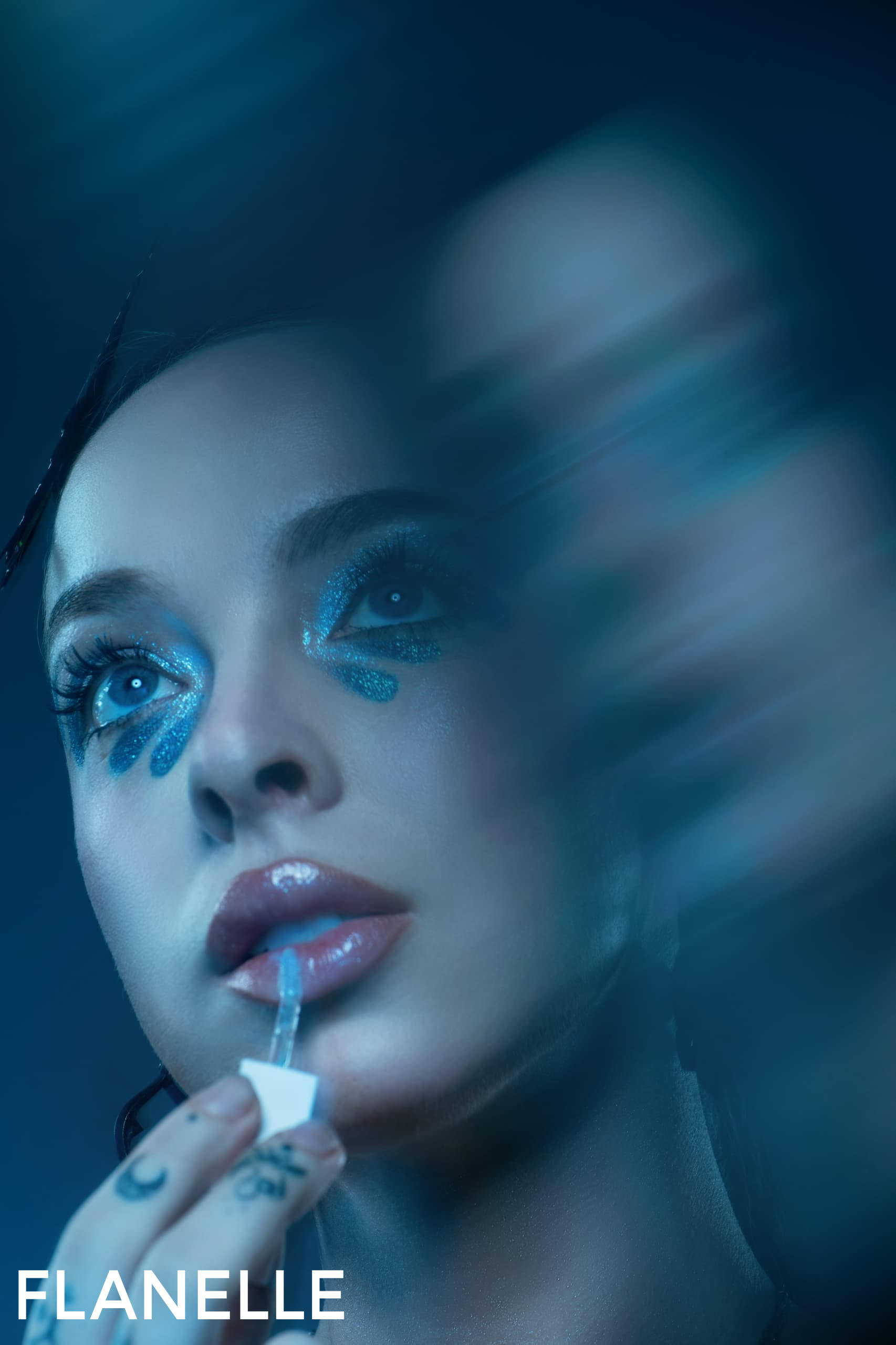 Woman applying lip gloss, featuring bold blue eye makeup, under dramatic lighting.