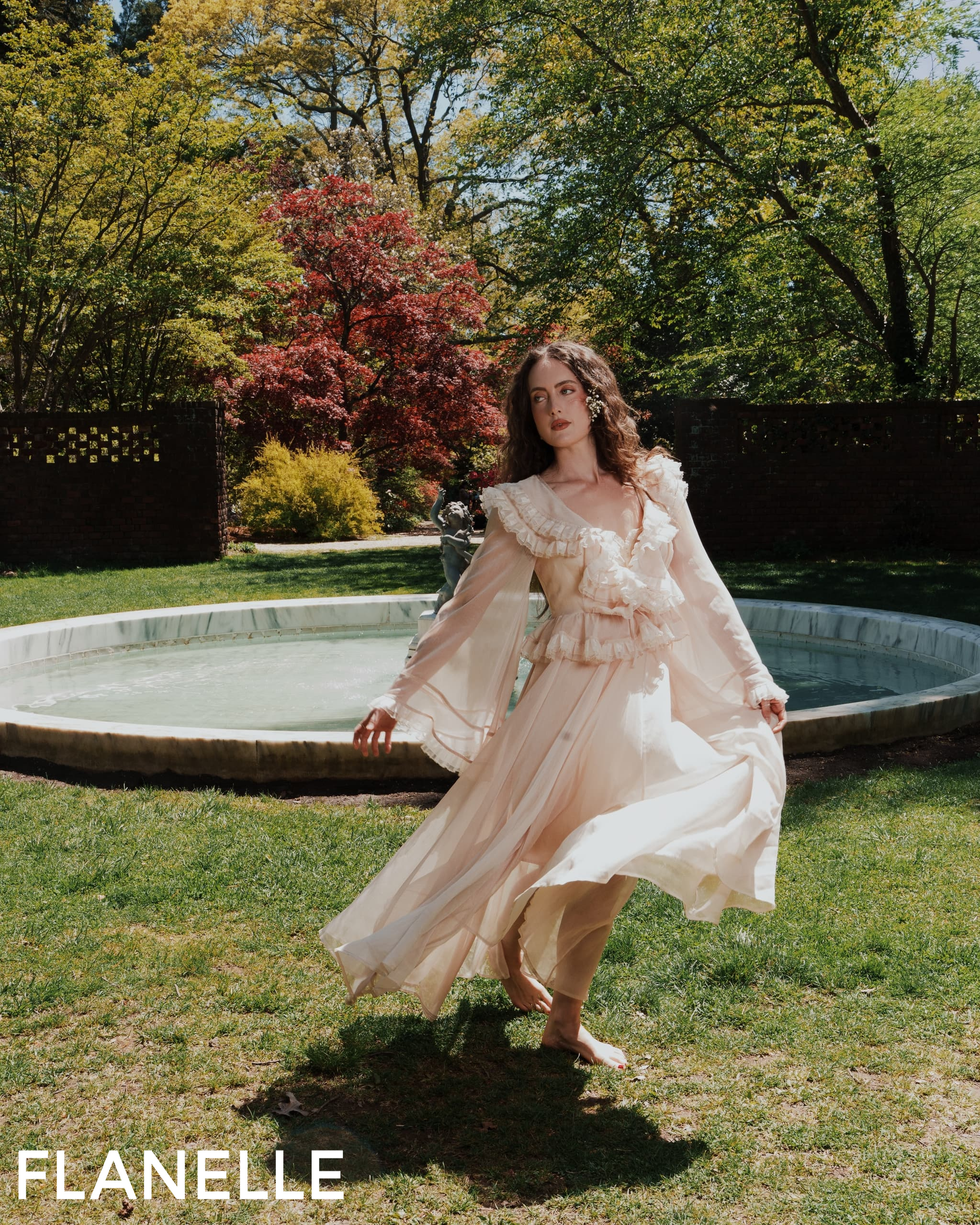 Woman in a flowing dress near a fountain in a lush garden.