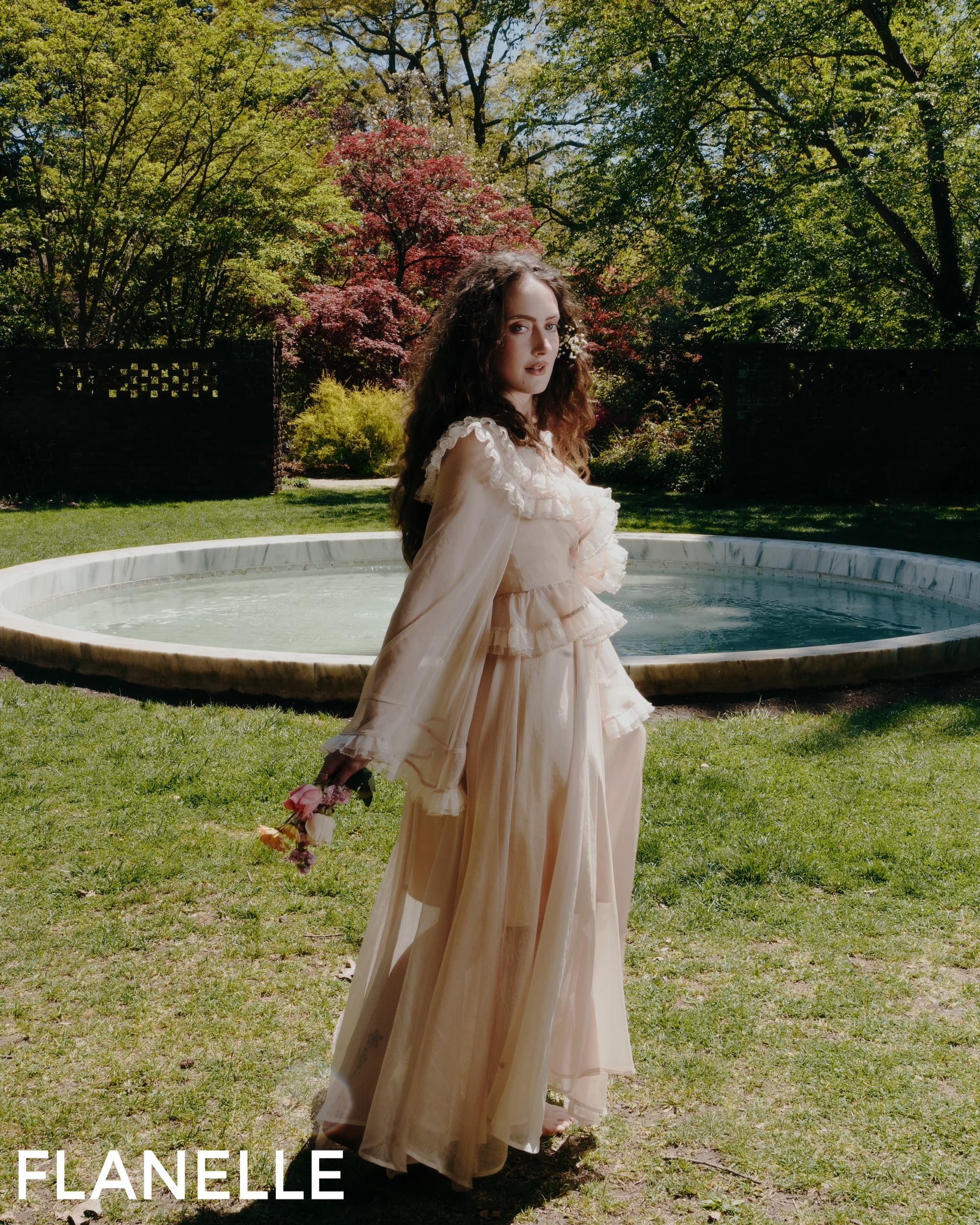 Woman in flowy dress by a garden pond, holding flowers, during spring day.