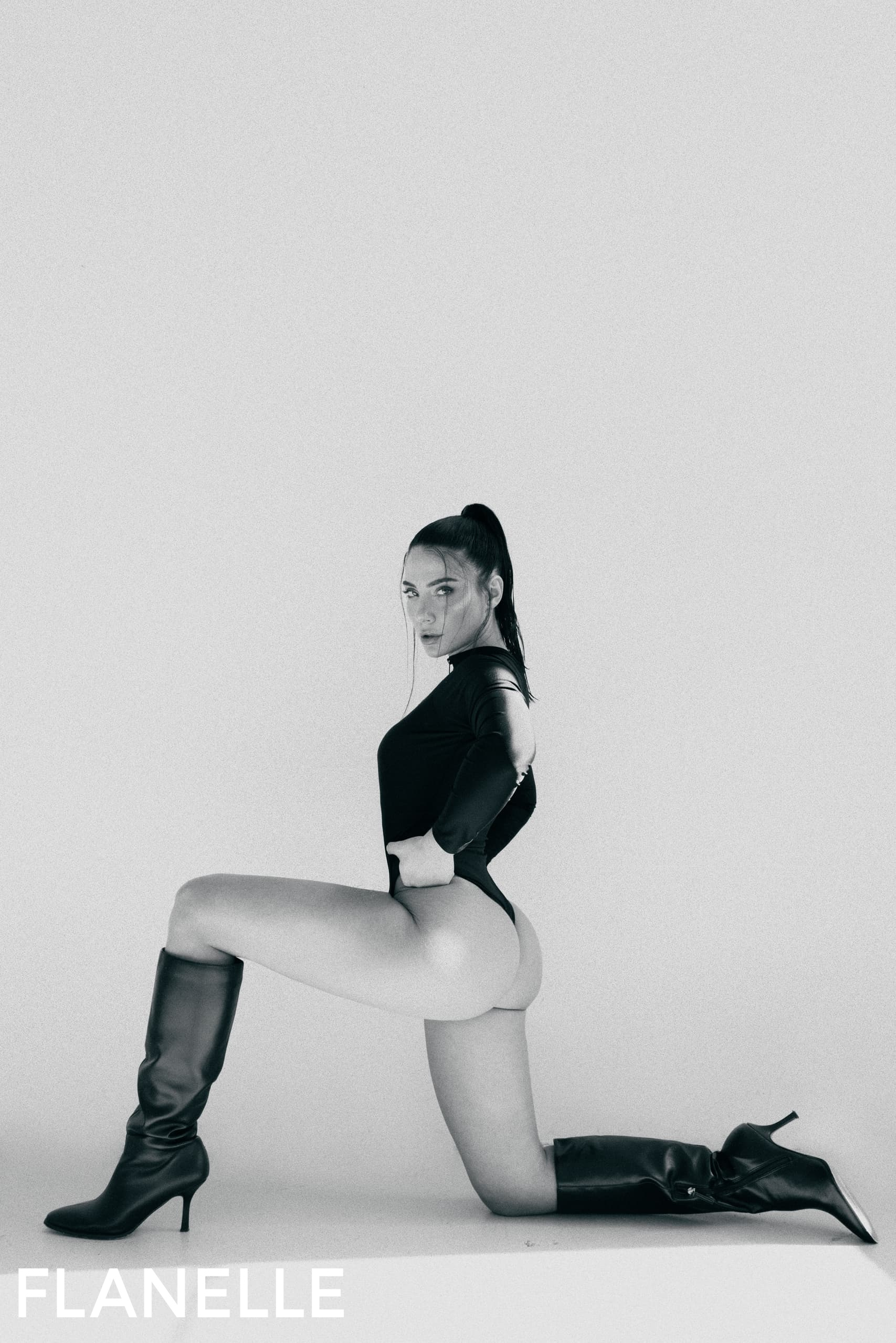 Woman in black bodysuit and boots posing in a minimal studio setting.