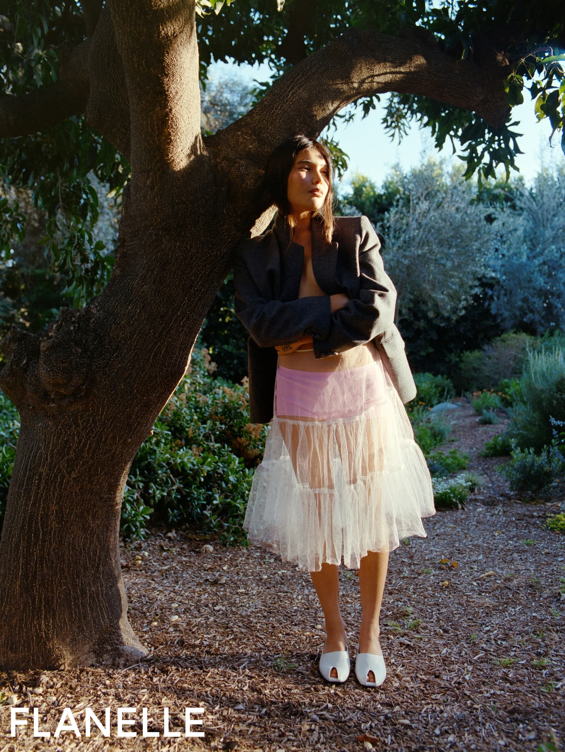 Fashion model in tulle skirt and blazer under tree in garden.