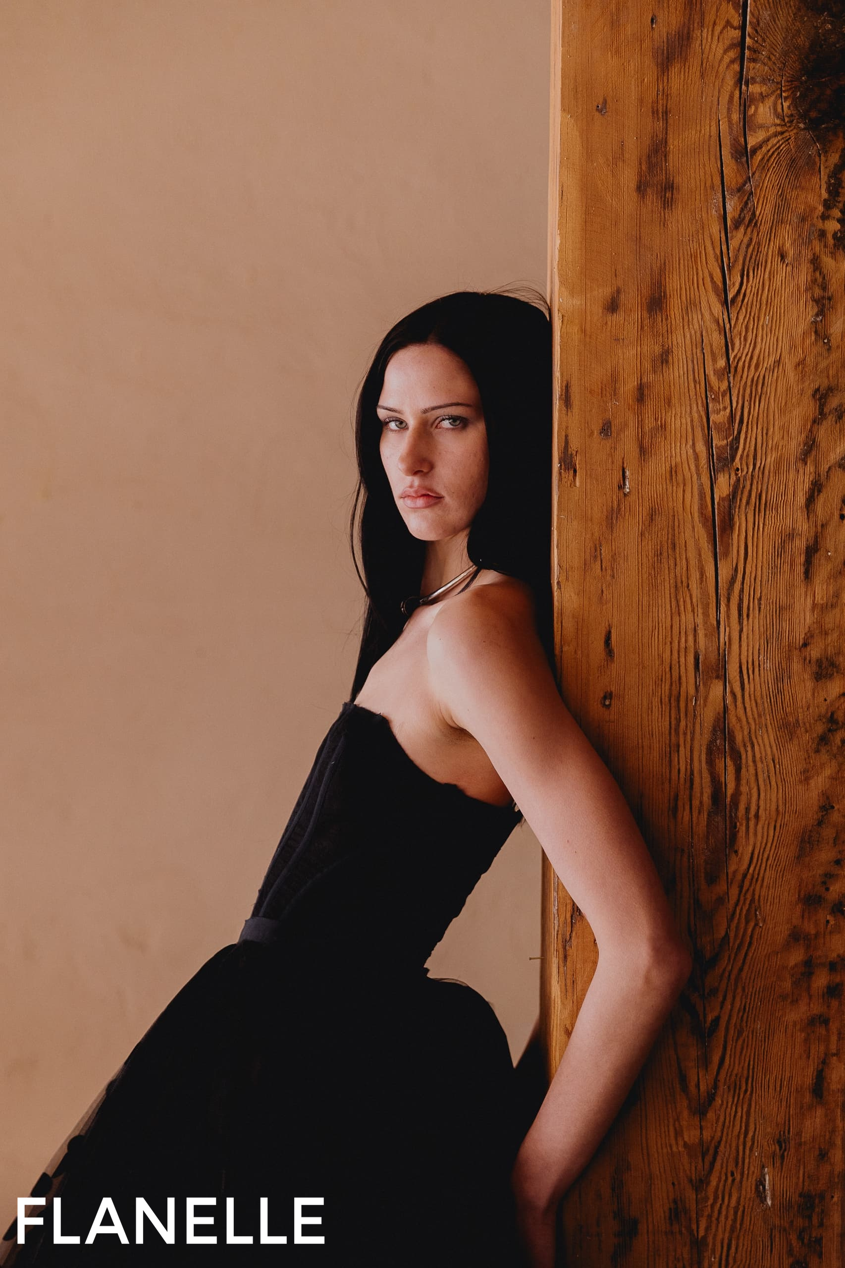 Fashion model in black dress leaning against a wooden post.