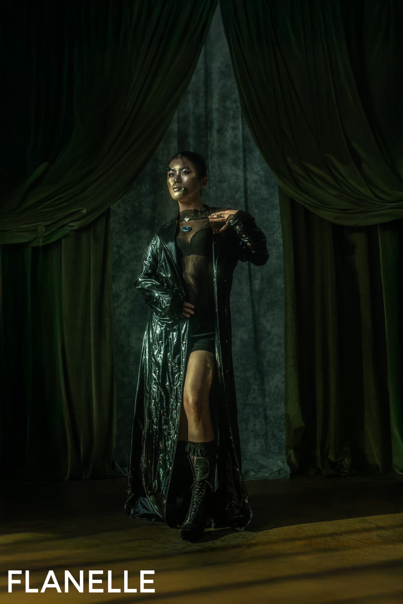 Model in black long coat poses with dramatic lighting and green curtains.
