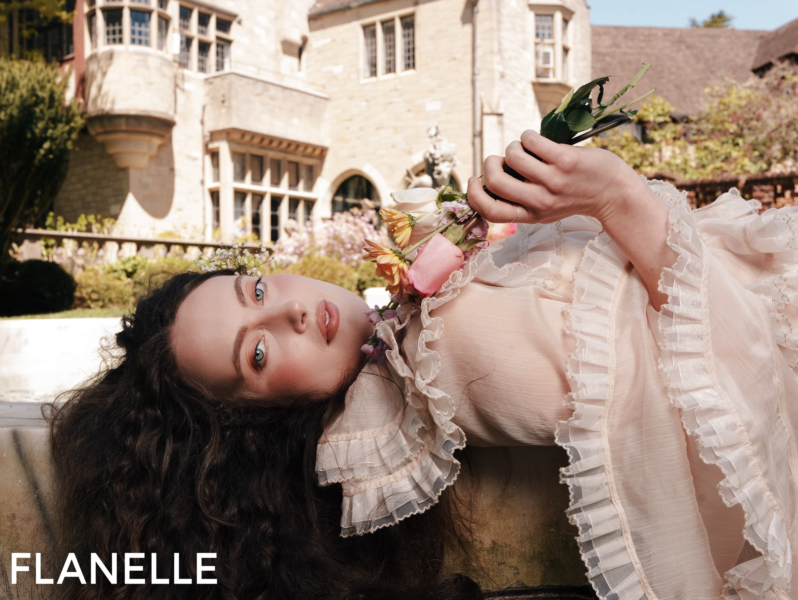 Woman in a ruffled dress lying outdoors, holding flowers near historic building.