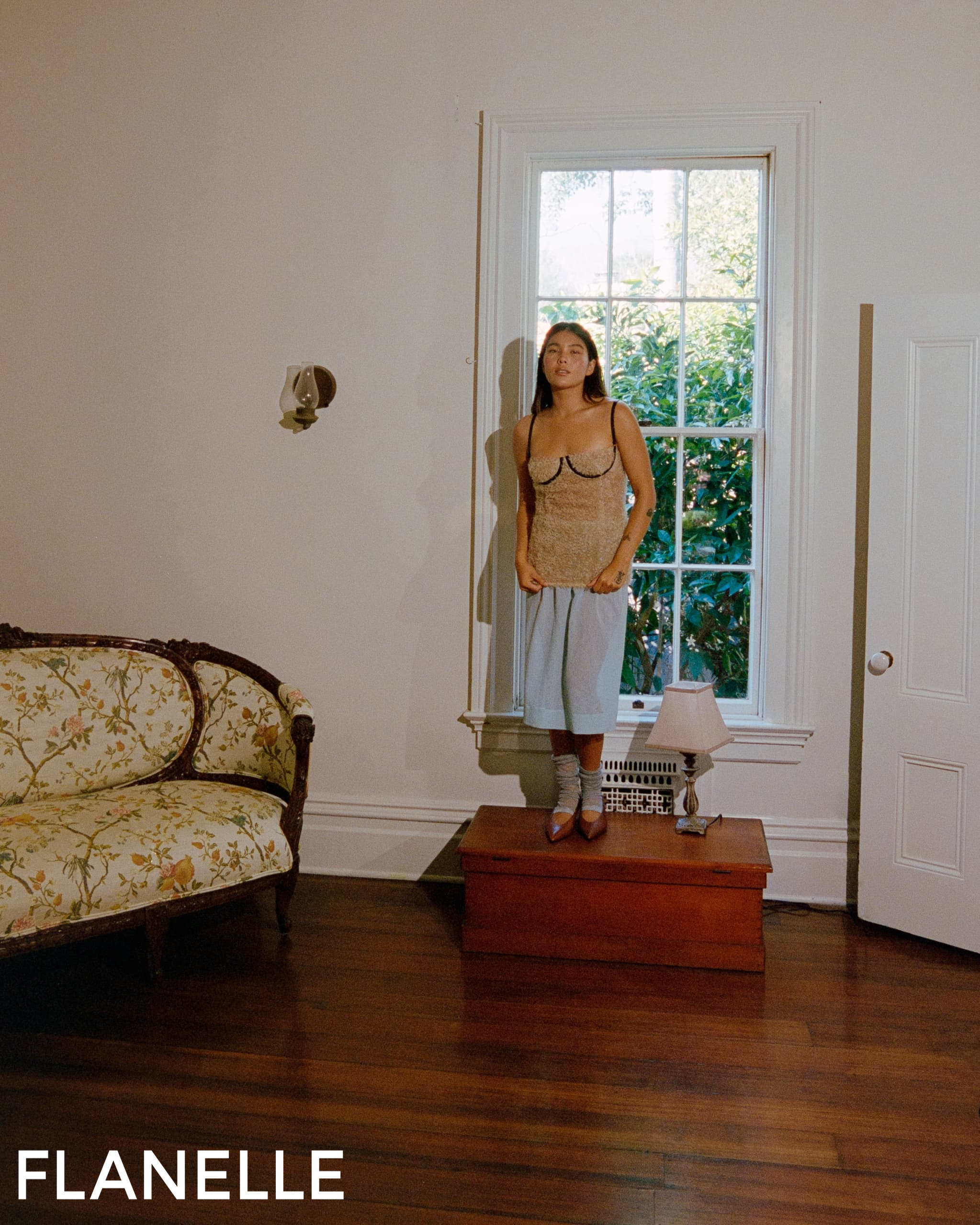Woman in stylish outfit stands by a window in a vintage room.