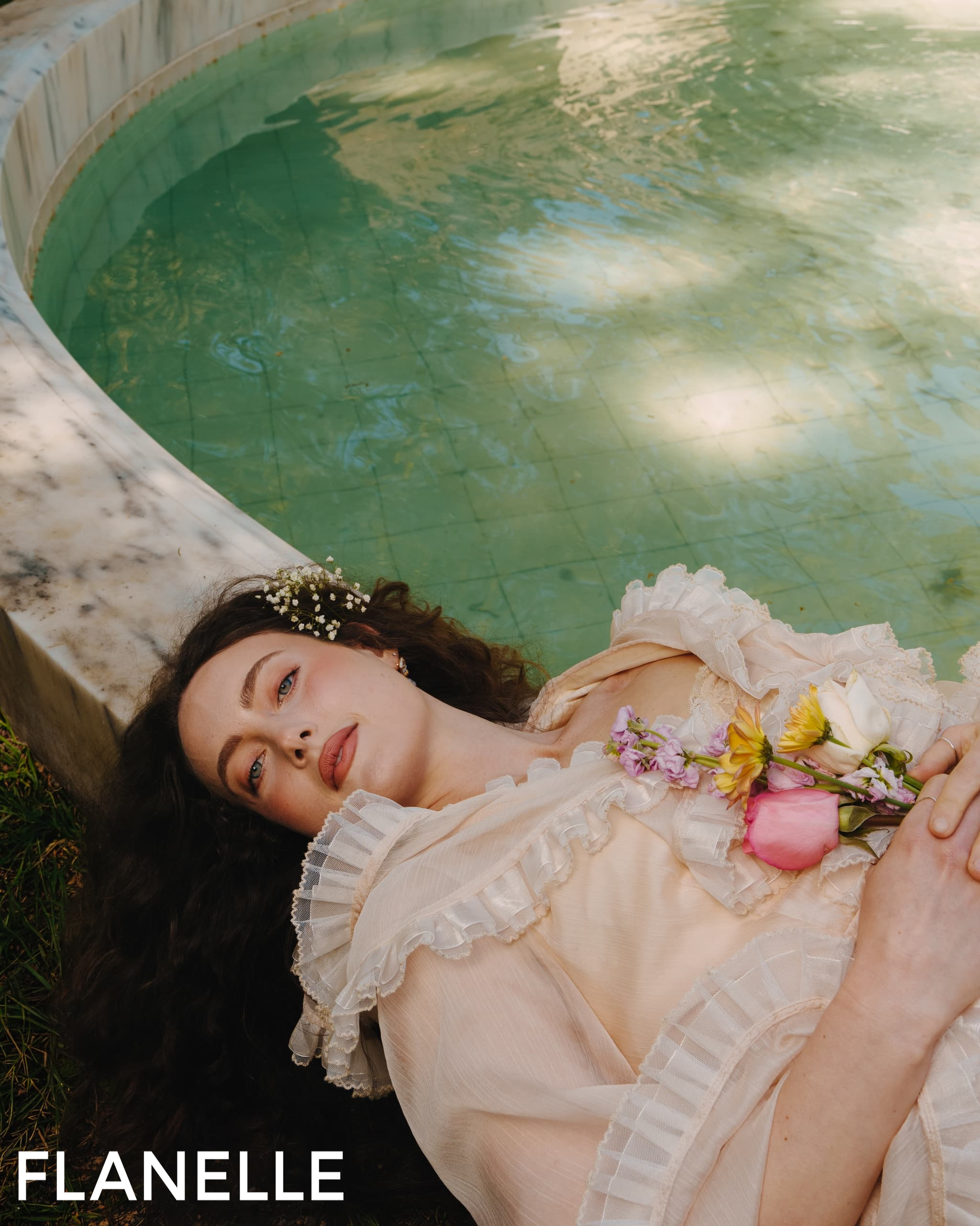 Woman in white dress with flowers, lying by pool. Romantic scene. Flanelle magazine.