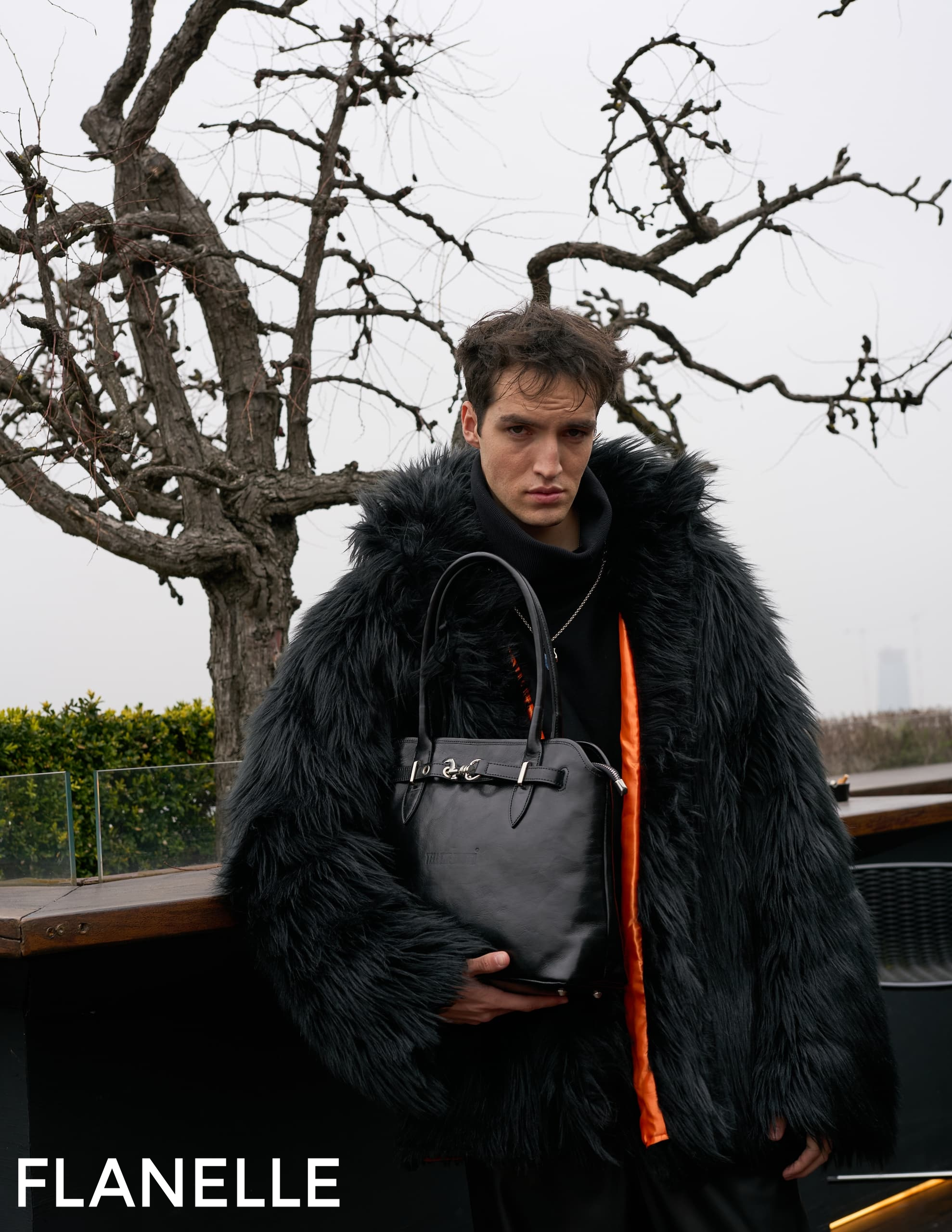 Man in black fur coat holds leather bag with barren winter trees in the background.