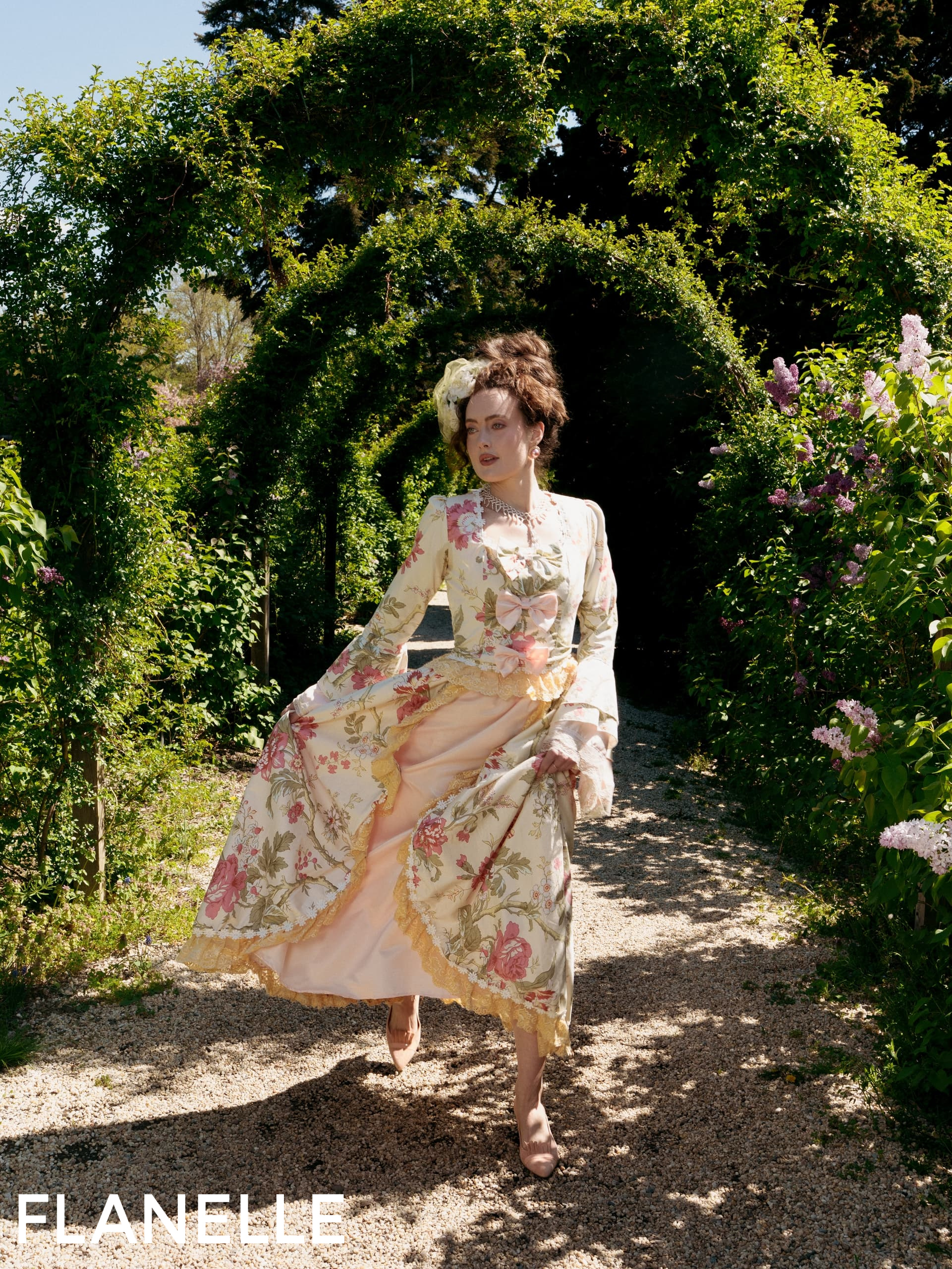 Elegant woman in floral gown walking through sunlit garden arches.