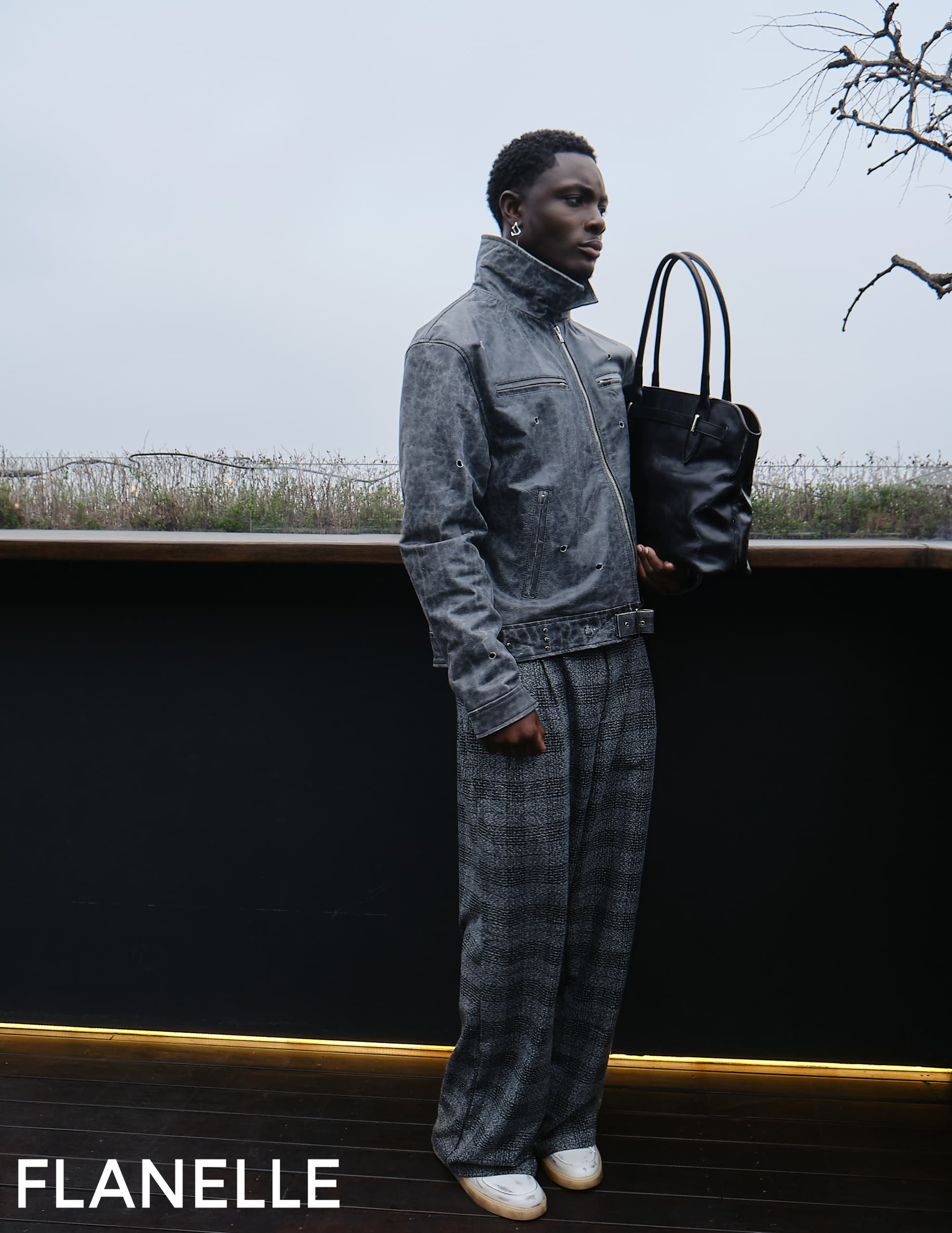 Fashionable person in gray outfit with black tote next to leafless tree.