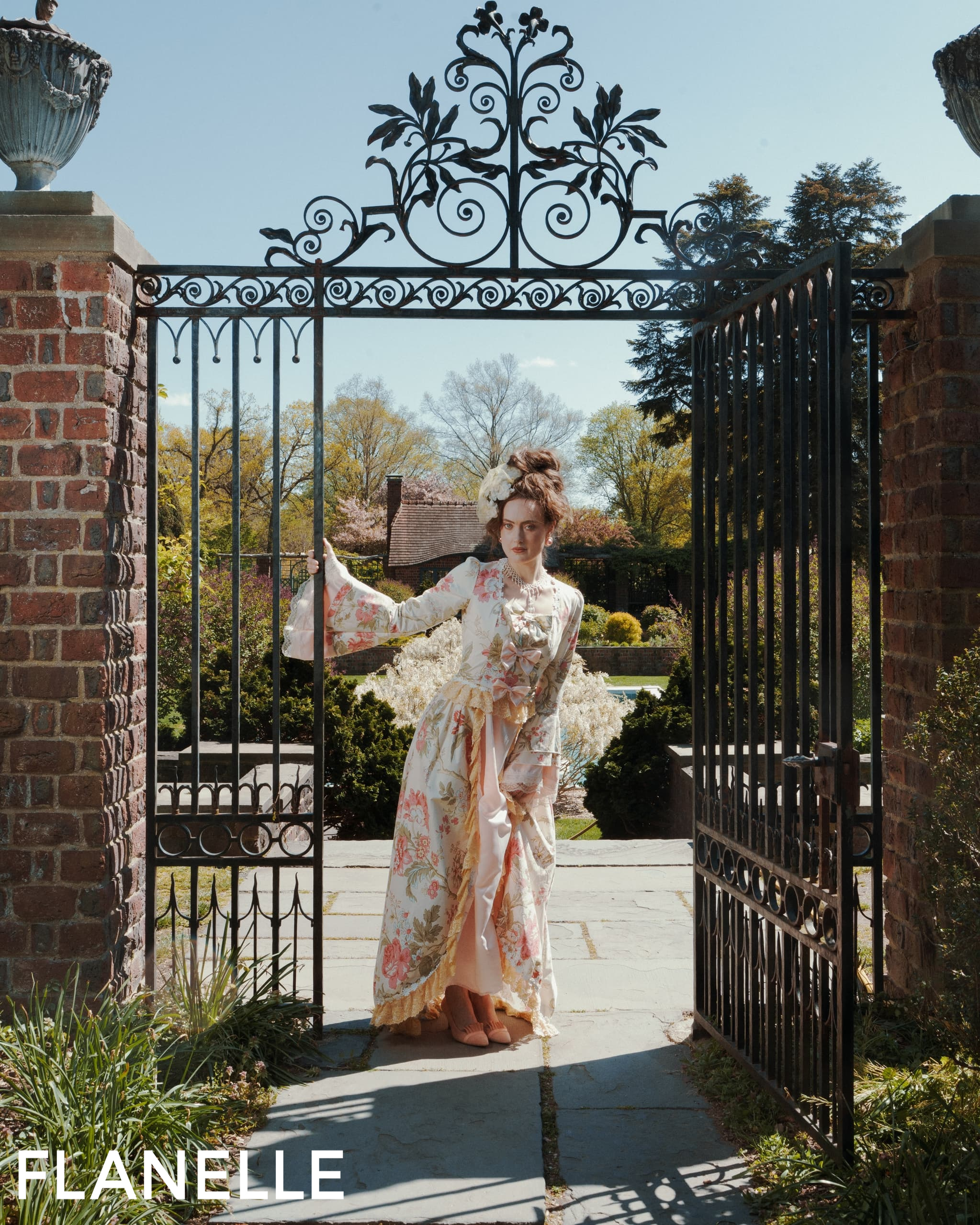 Woman in floral dress stands by ornate garden gate.