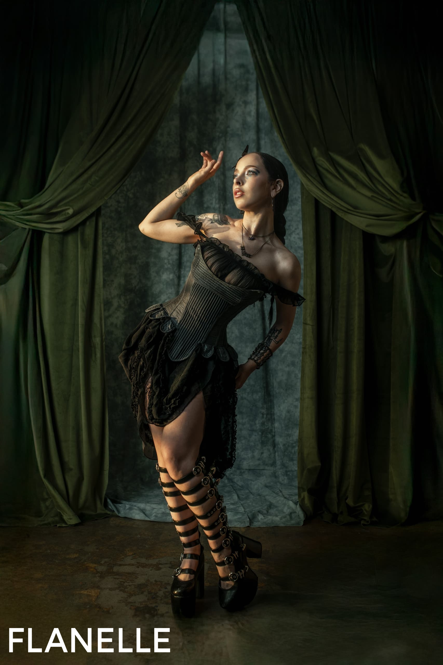 Gothic fashion model poses in black corset dress against dark green curtains.