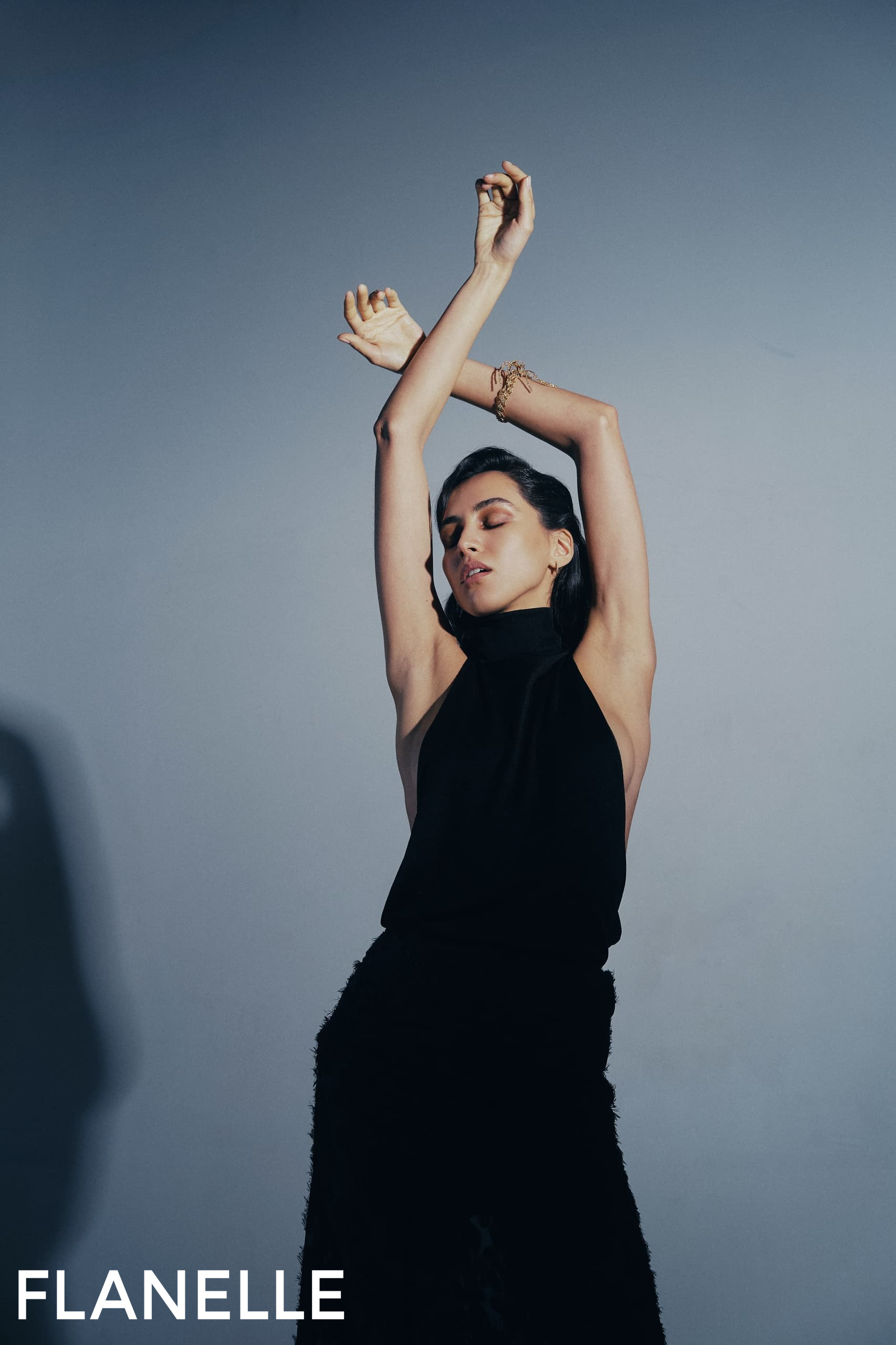 Fashion model posing with arms raised against a gray background in a black outfit.
