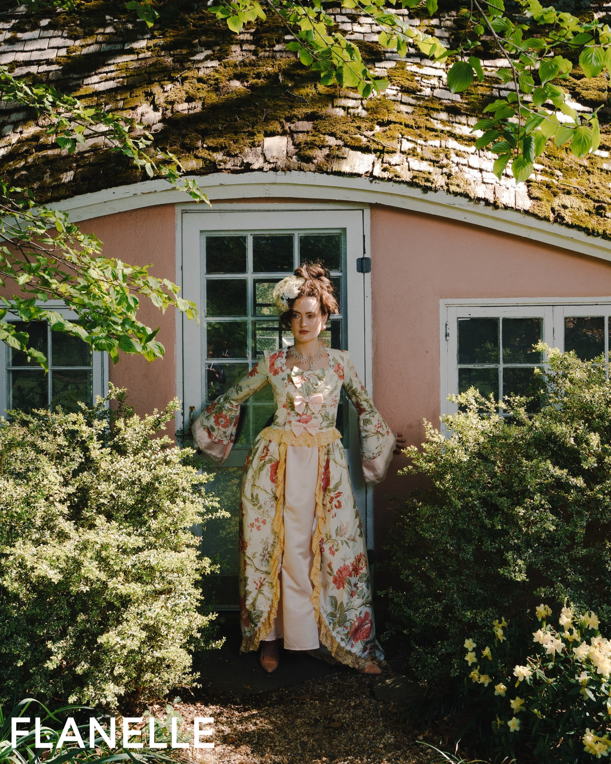 Woman in ornate floral dress stands by a rustic cottage door.