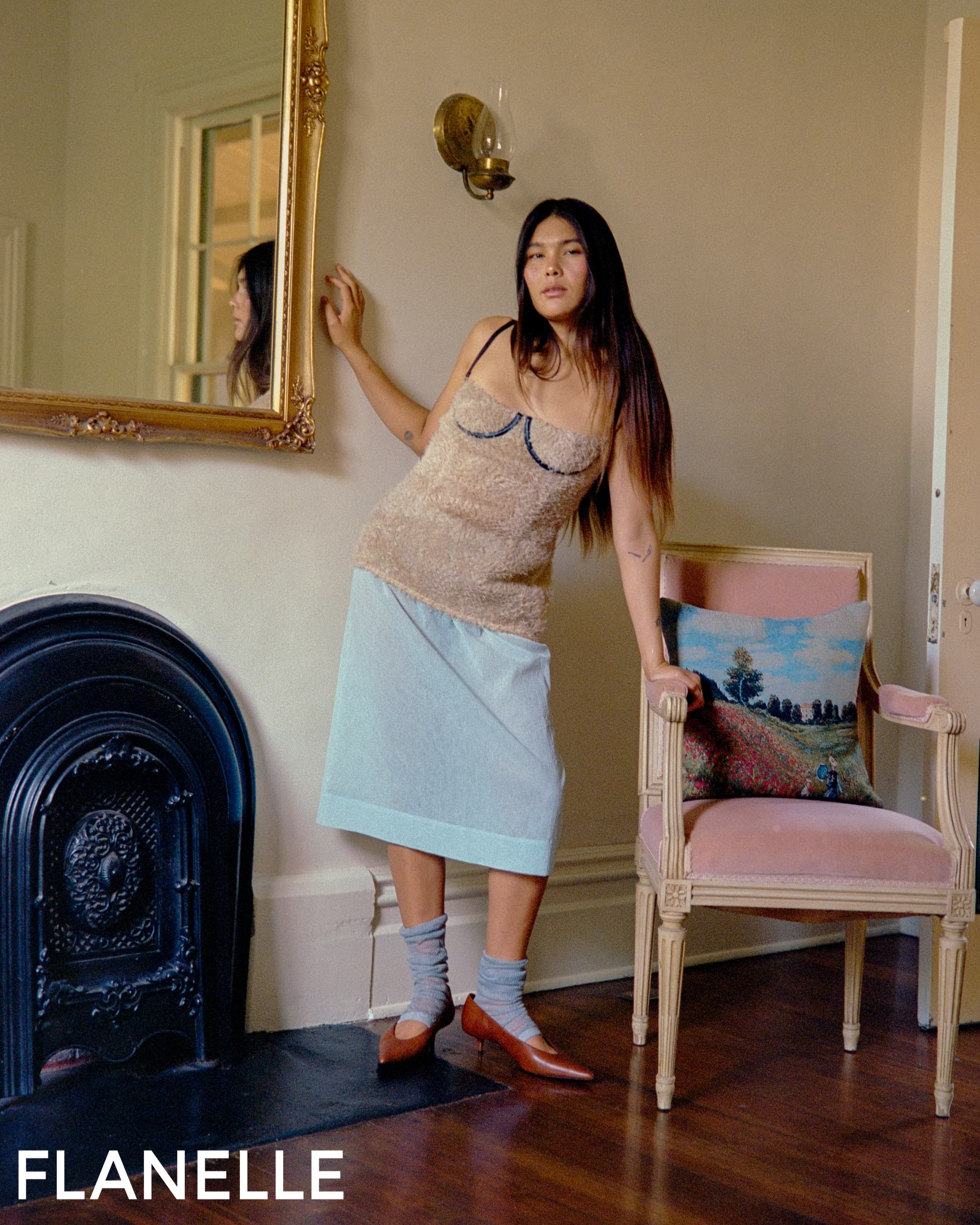 Woman in stylish outfit poses by ornate mirror and chair in vintage room setting.