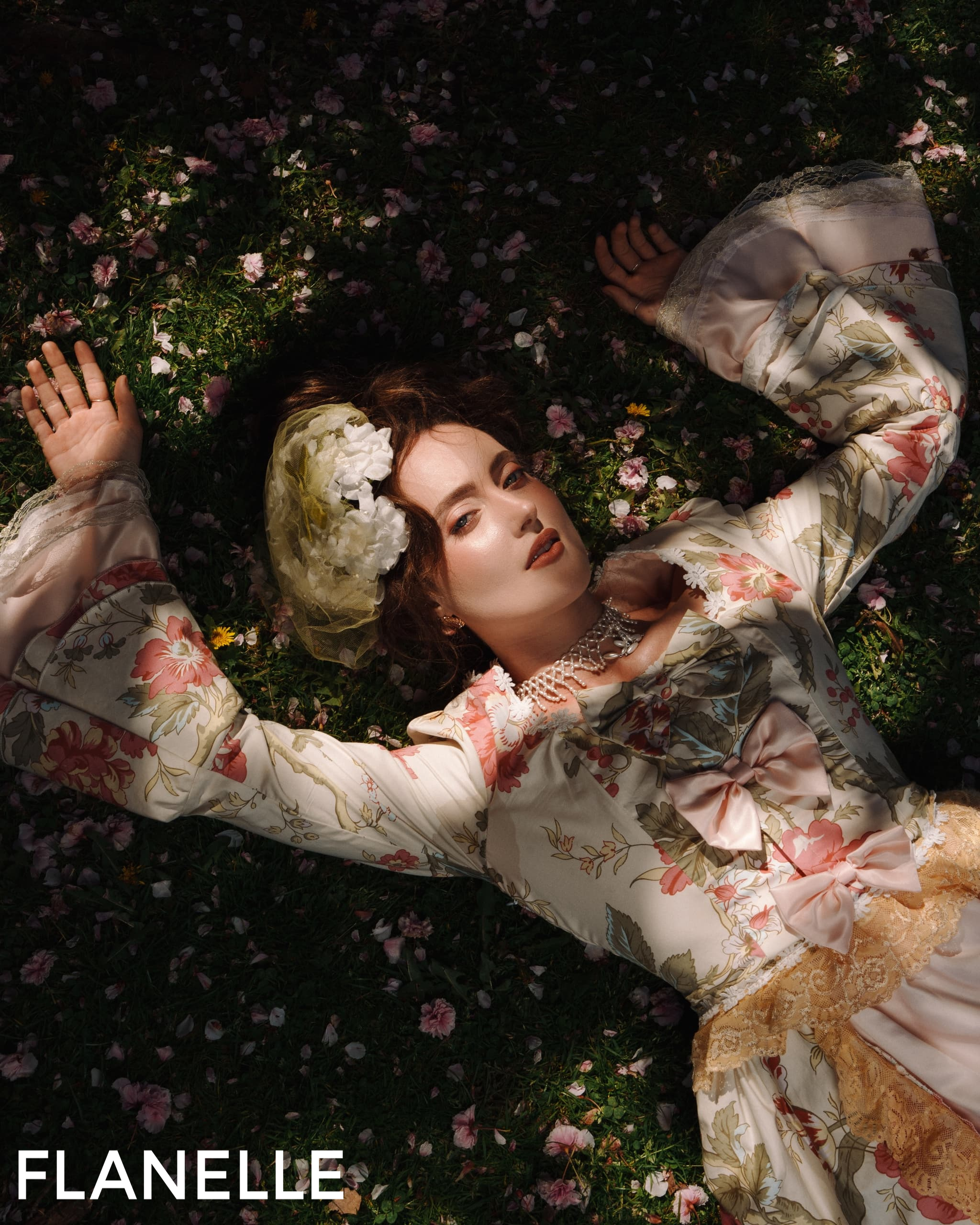 Woman in floral vintage dress lying on grass amidst scattered blossoms.