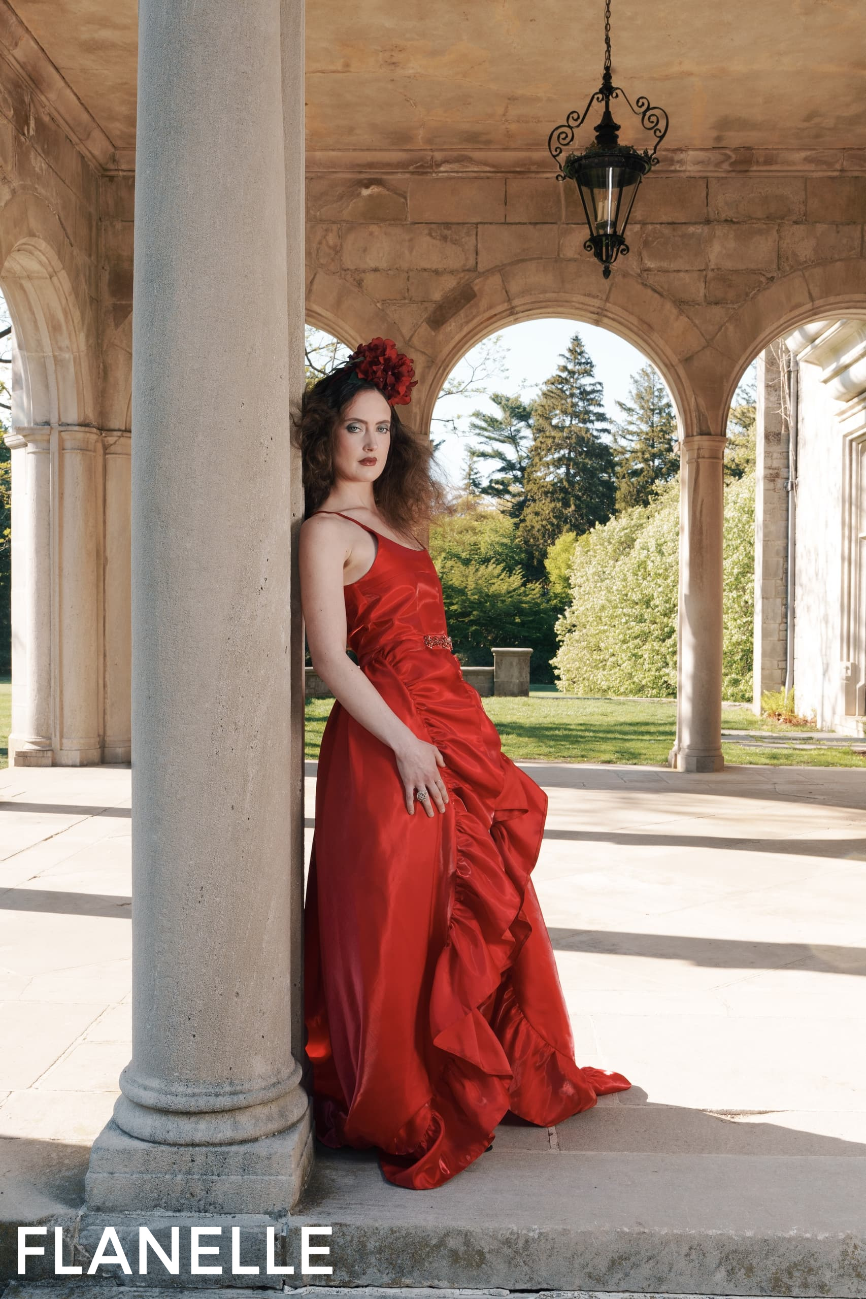 Woman in red dress leans against pillar in elegant garden setting.