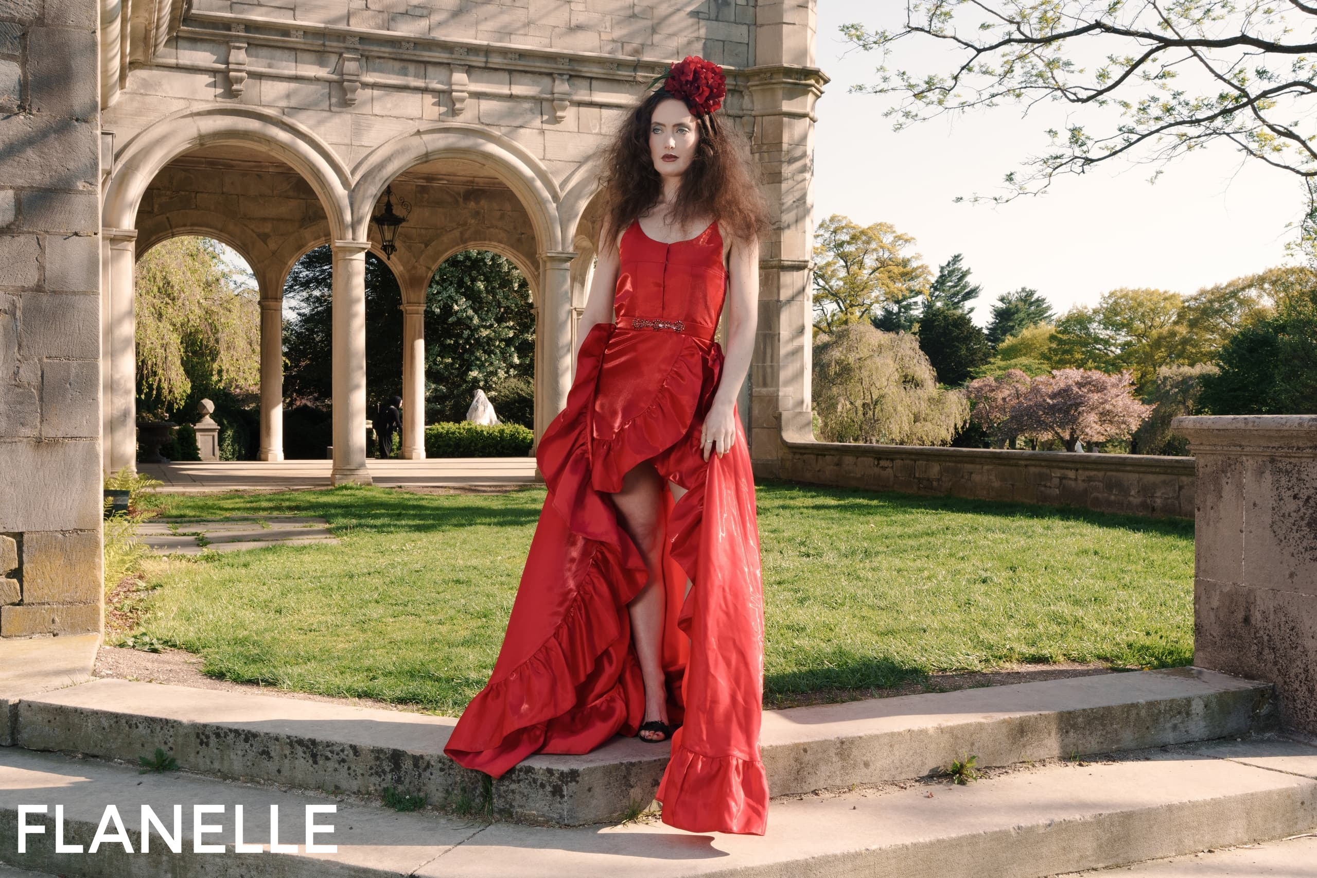 Woman in elegant red dress by stone arches, outdoor setting, sunny day.