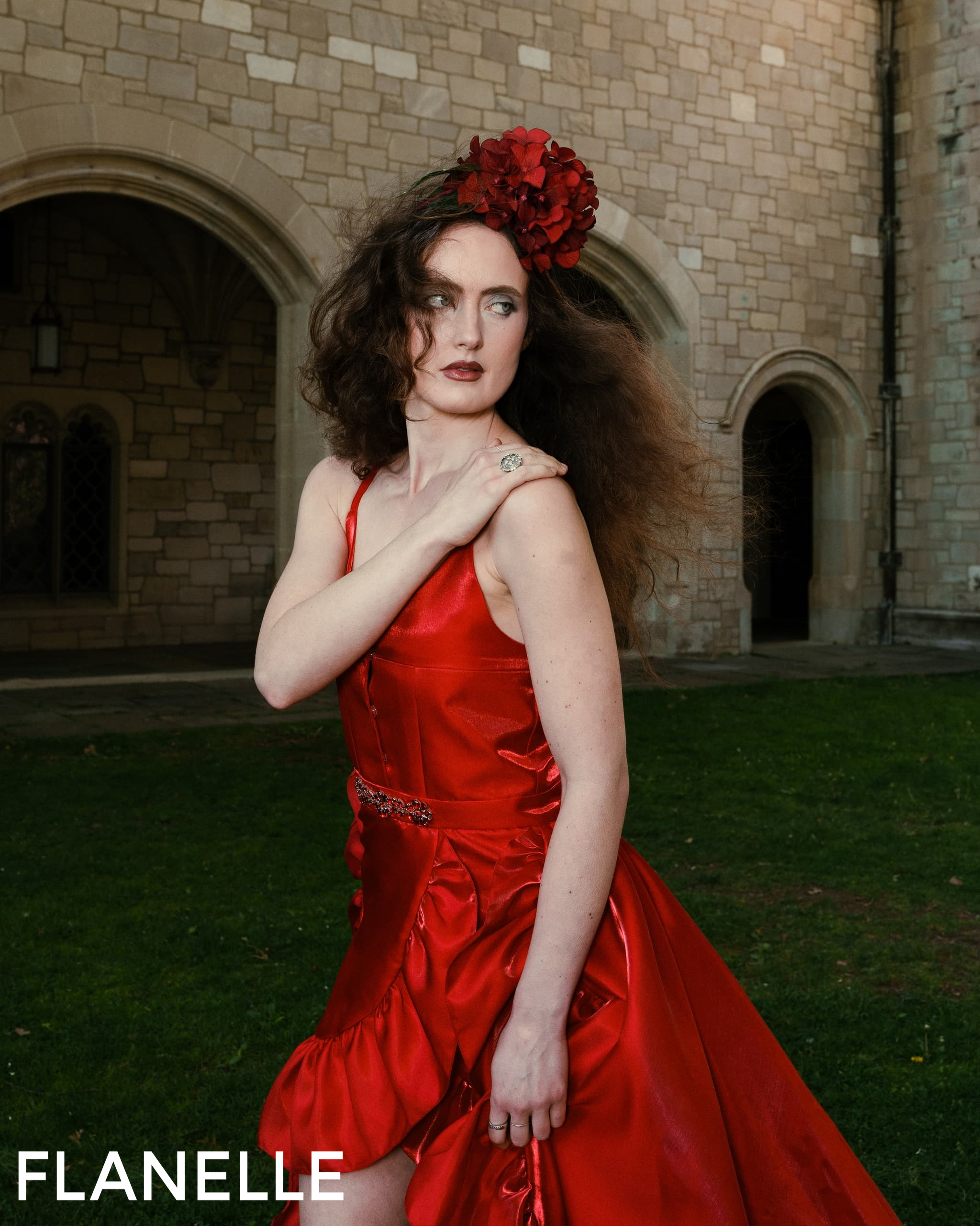 Woman in a red dress with red floral headpiece near stone building.