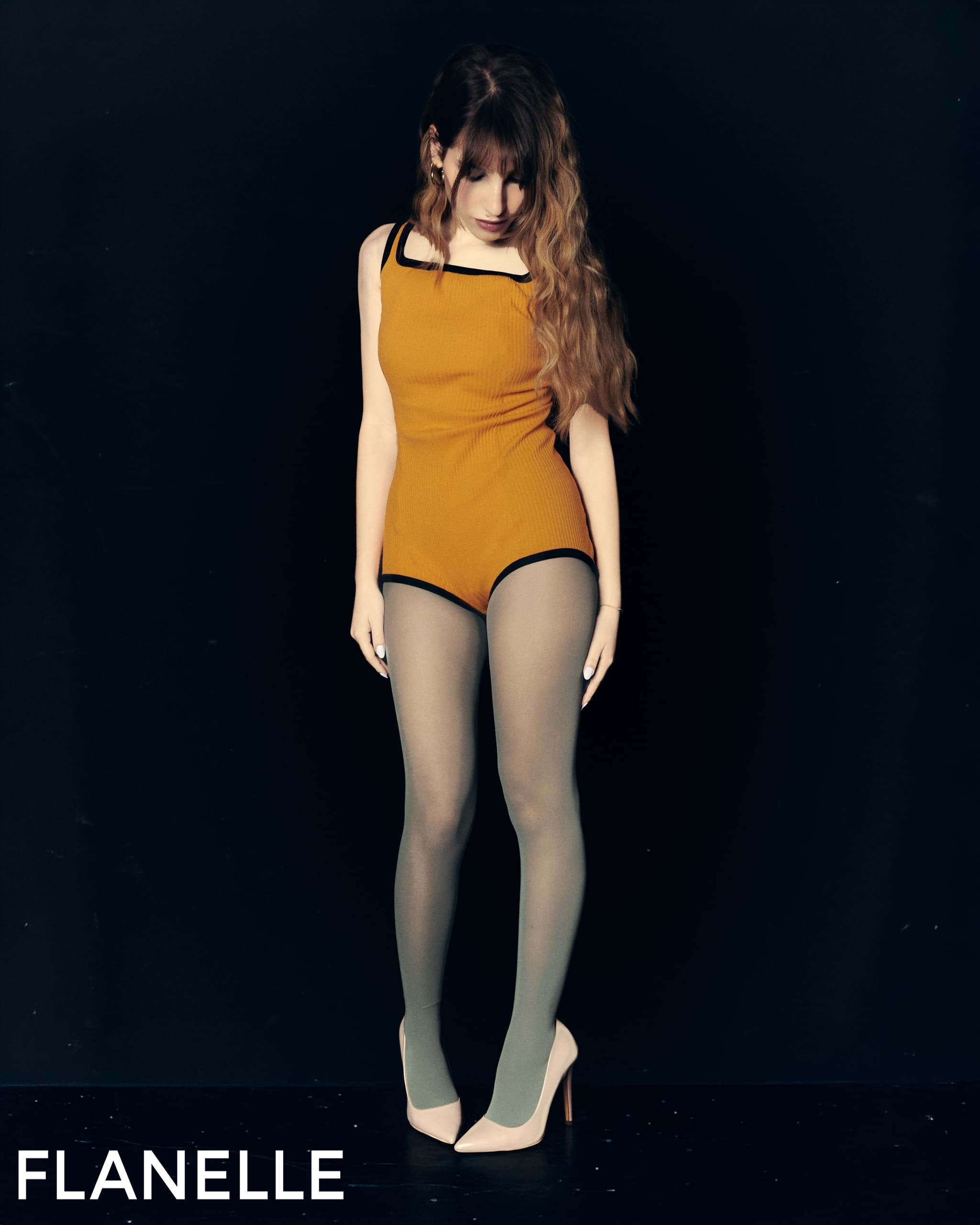 Fashion model in an orange outfit with tights and heels against a dark background.