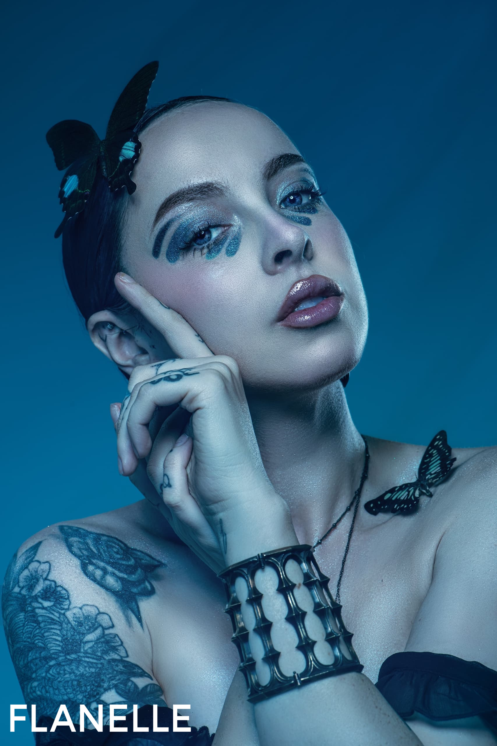 Woman with butterfly makeup and tattoos in an artistic blue-toned portrait.