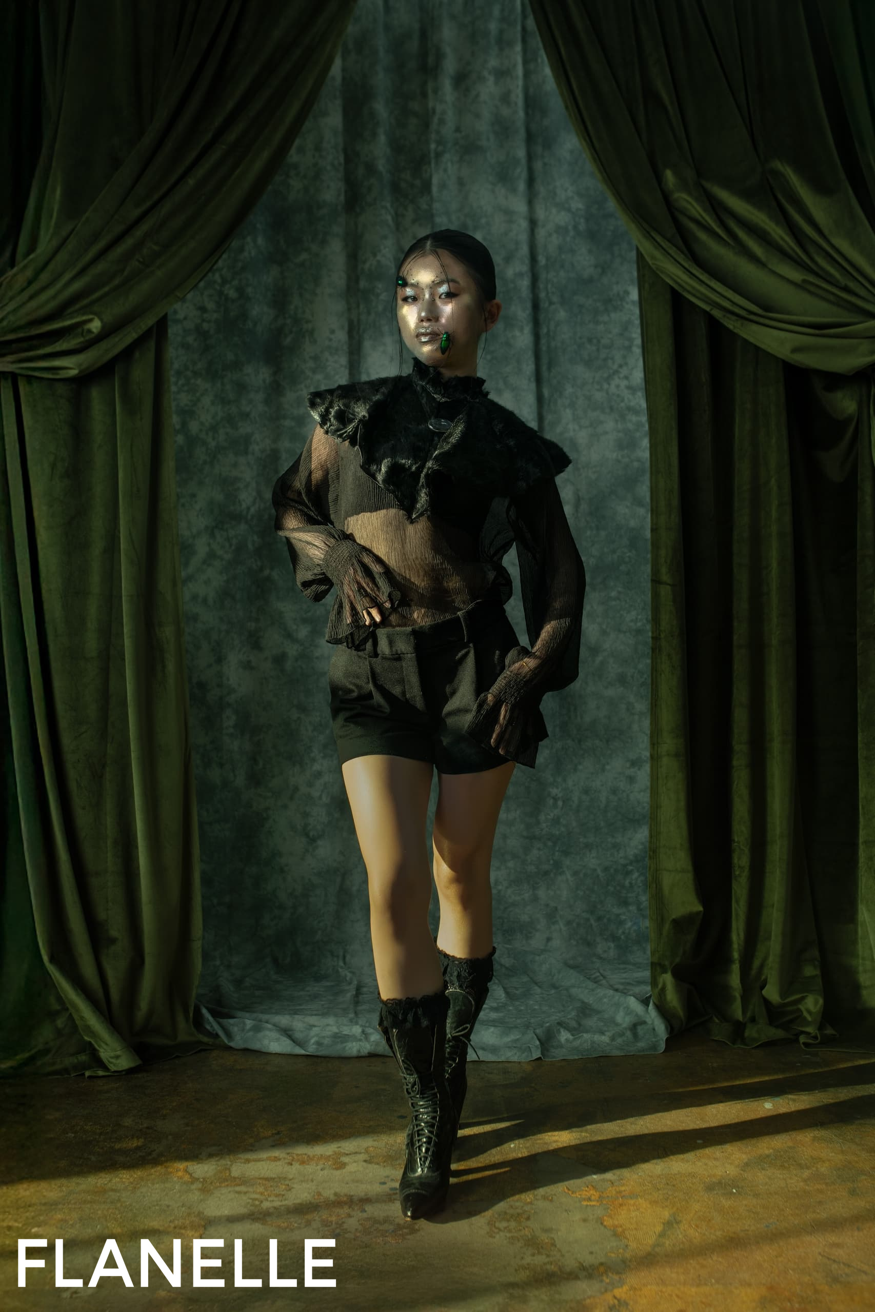 Fashion model in dark, stylish outfit on moody green backdrop.