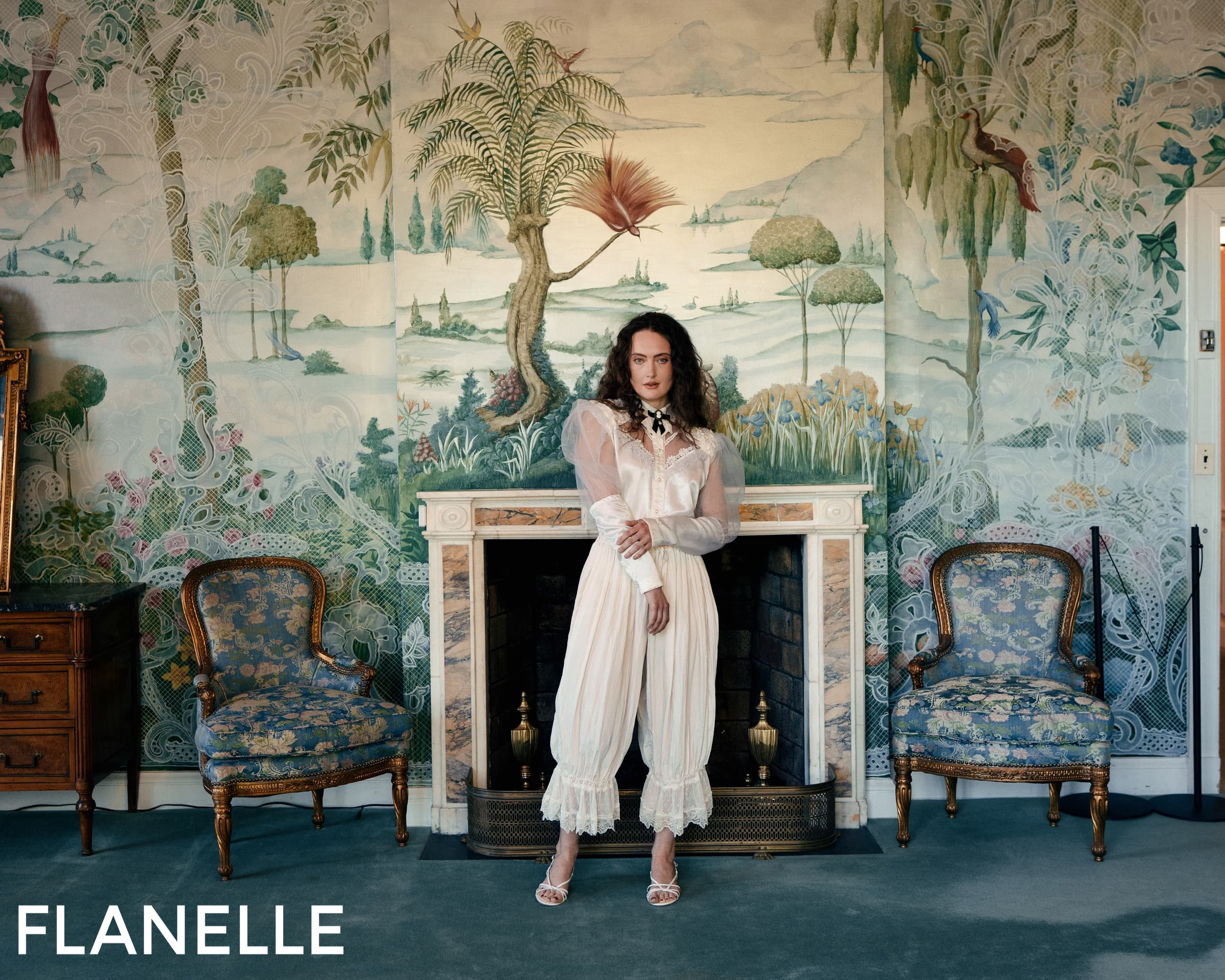 Woman in elegant white dress posing in vintage-themed room with scenic wallpaper.