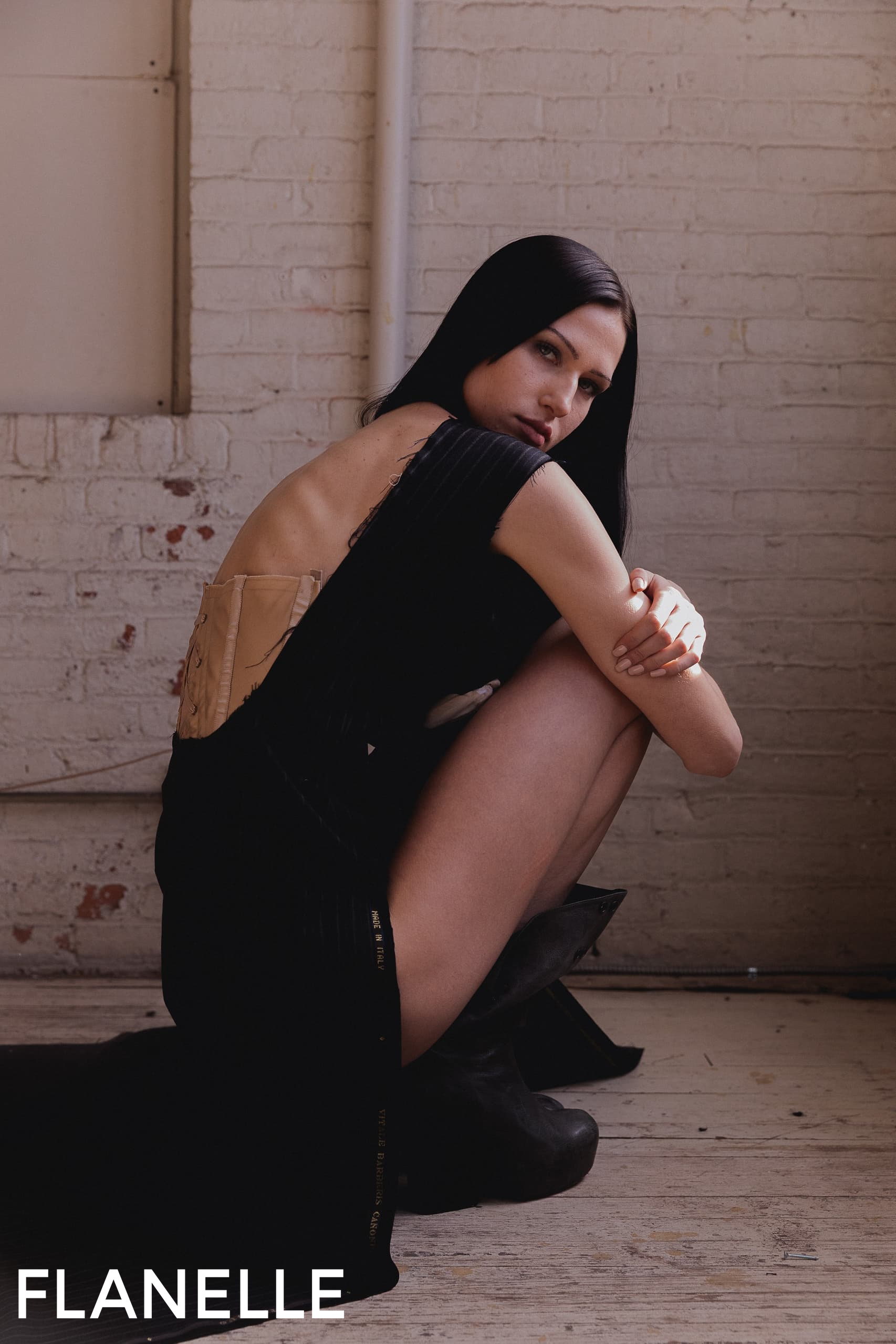 Model in a stylish backless black dress kneeling on a wooden floor.