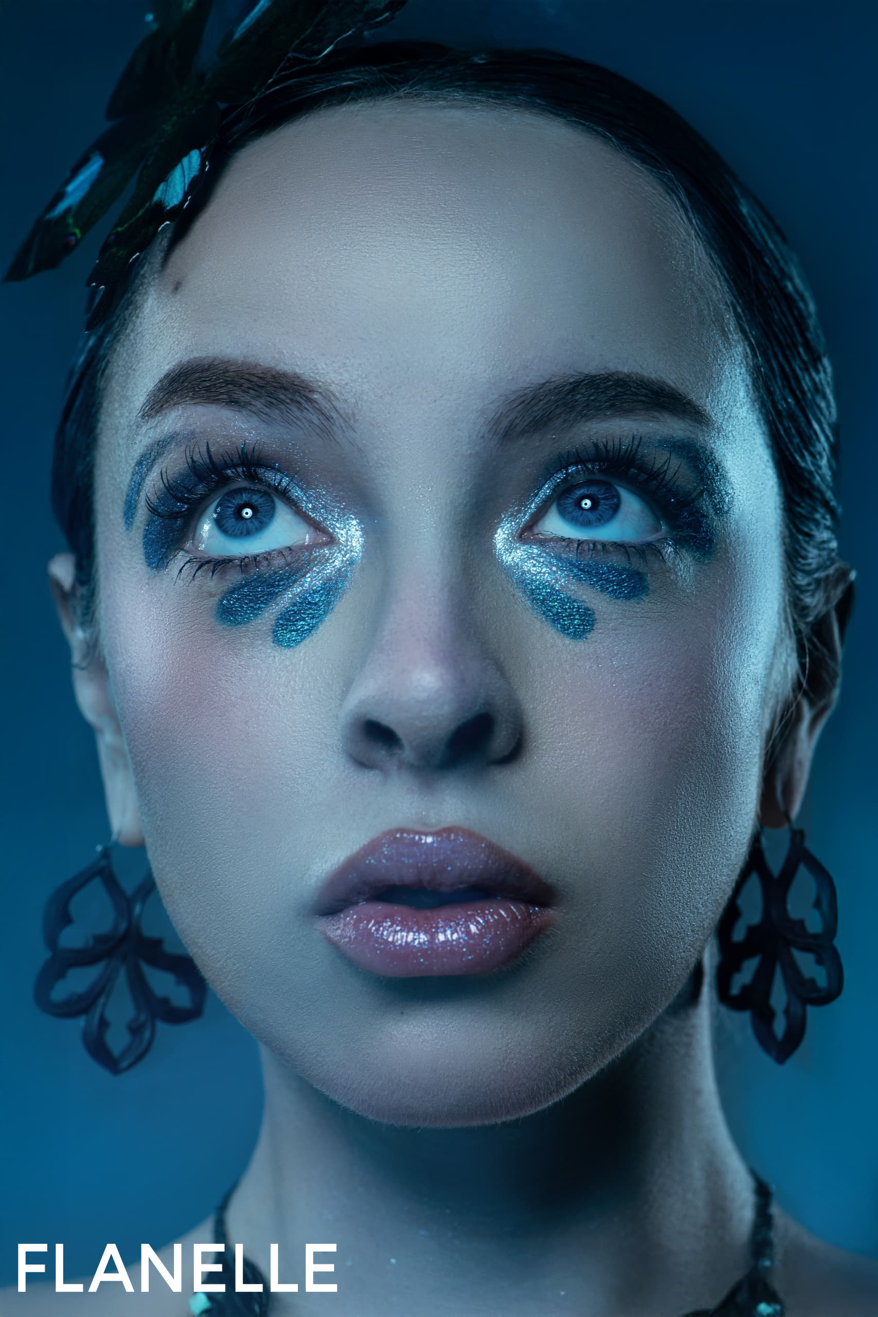 Woman with blue makeup and earrings, looking upwards in a moody blue tone.